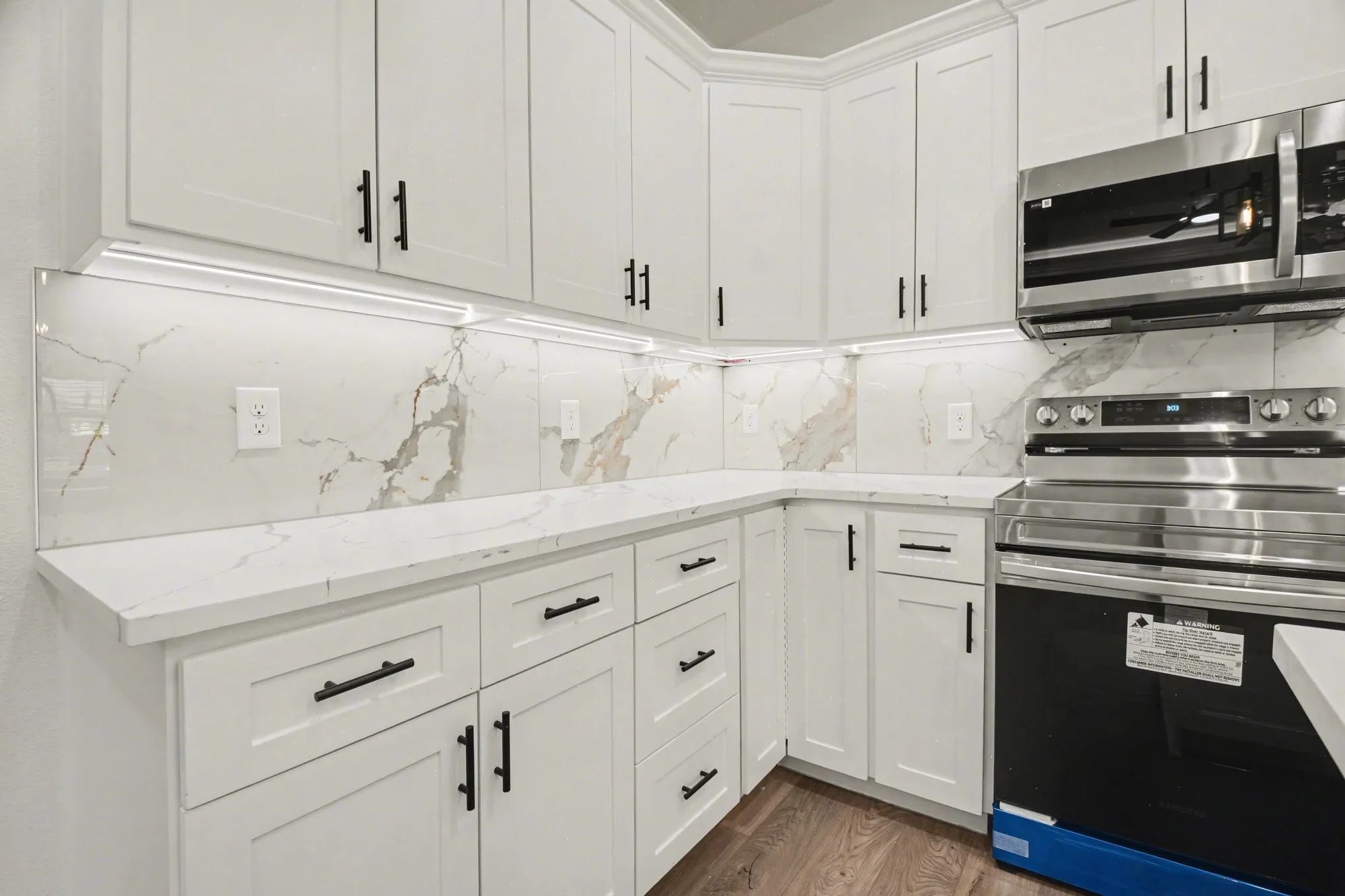White cabinetry with black hardware, stone-look countertops, matching backsplash, stainless steel appliances, and wood-finish flooring