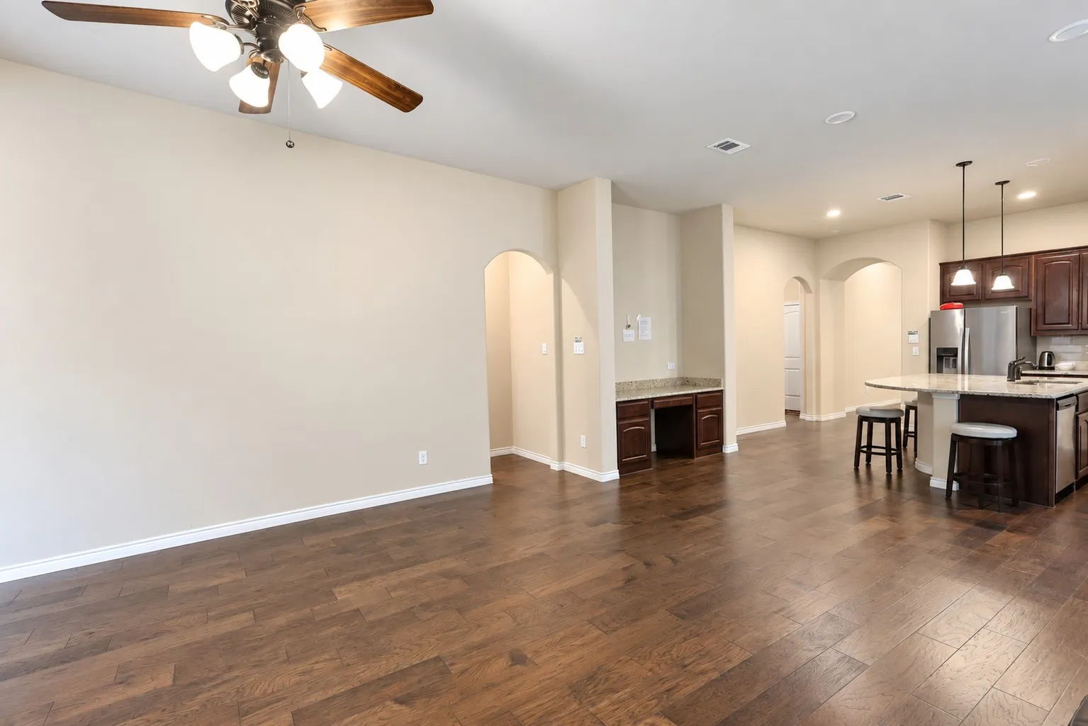 Open-concept living space featuring wood-finish flooring, a built-in desk area with granite-style countertop, a kitchen island with seating, pendant lighting, and arched doorways