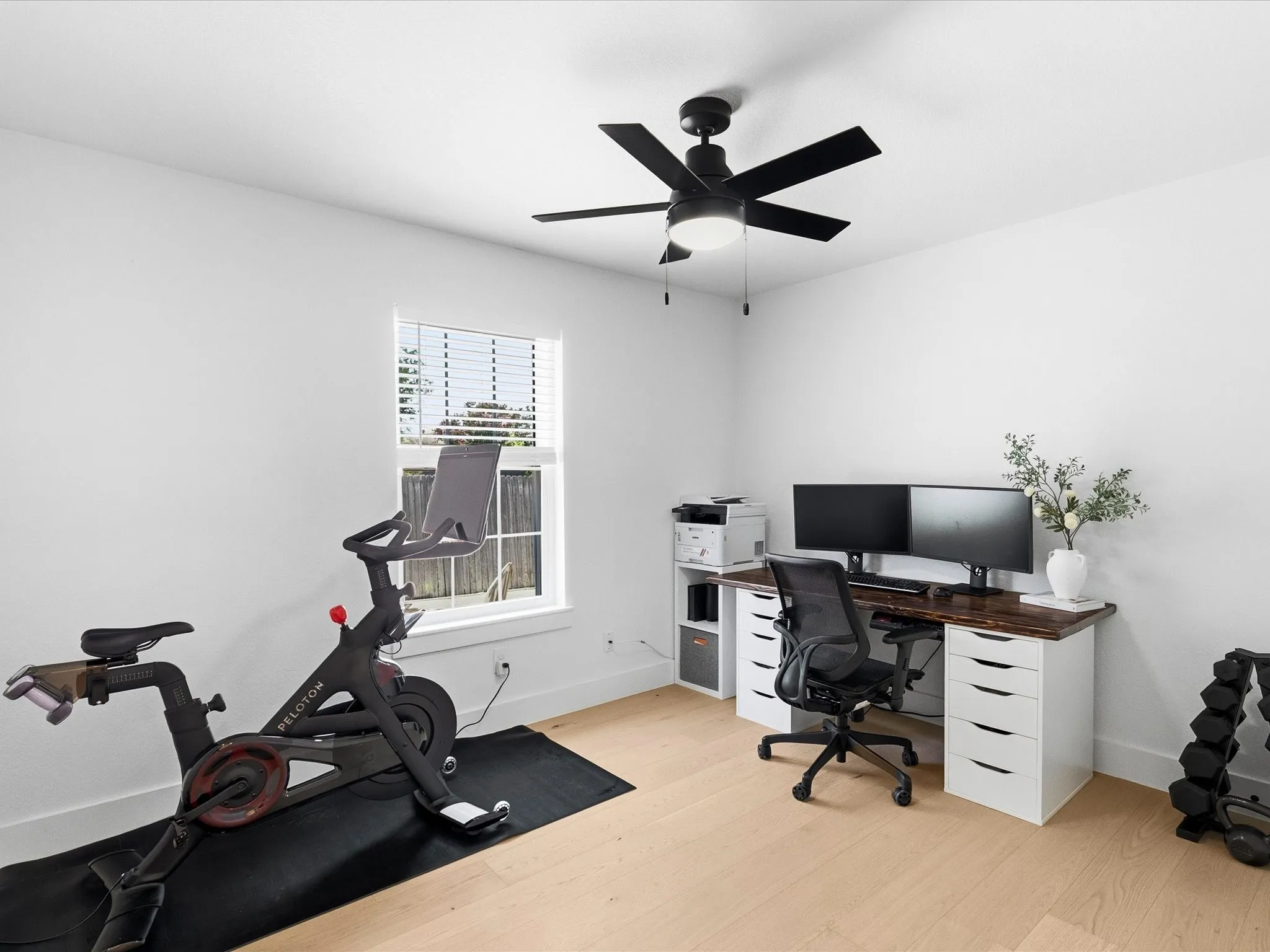 Light wood-finish flooring, white walls, rectangular window with blinds, and a black ceiling fan