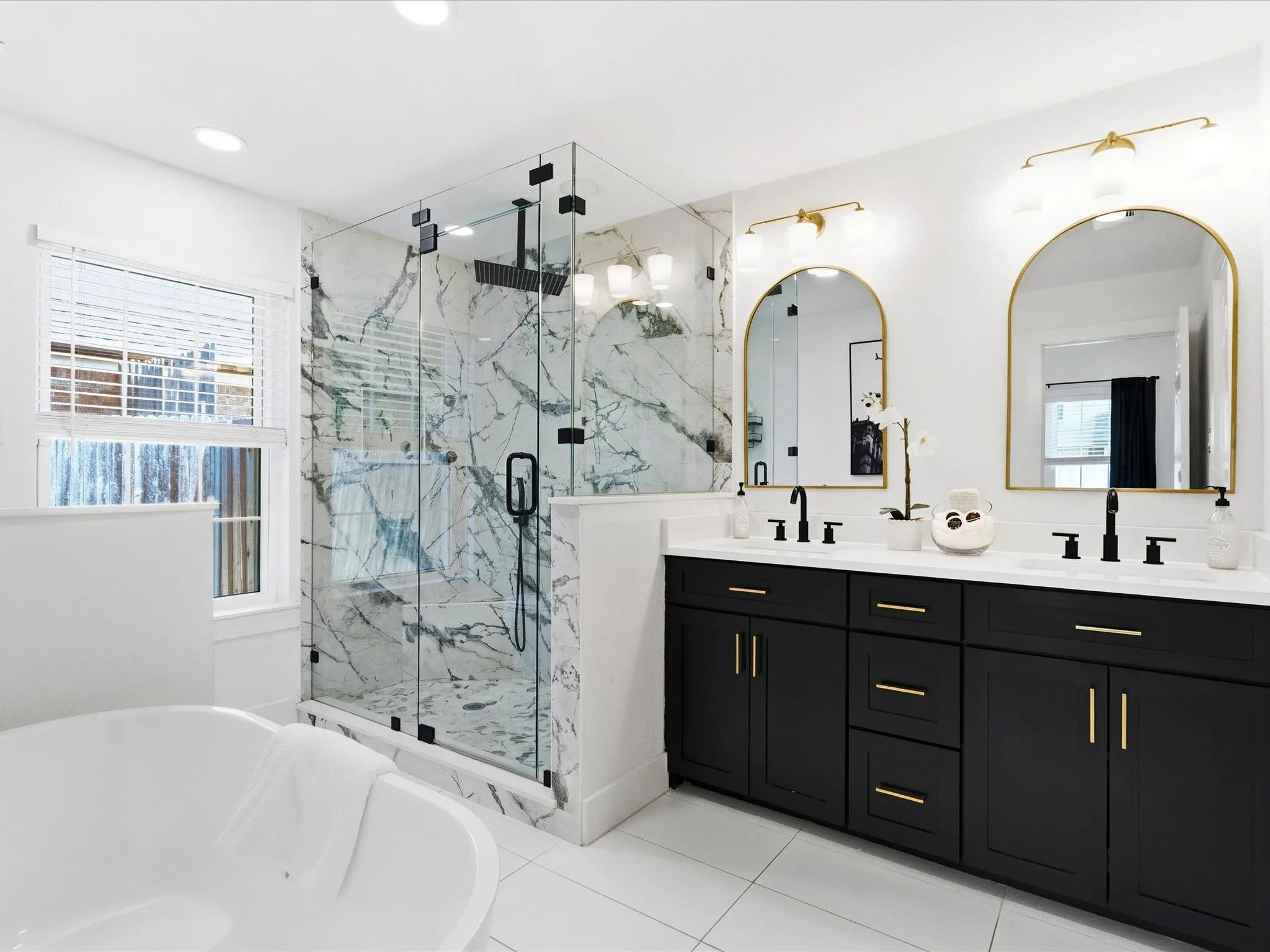 Freestanding bathtub, expansive walk-in shower with glass enclosure, dual vanity with black cabinetry, white countertops, and gold-framed arched mirrors