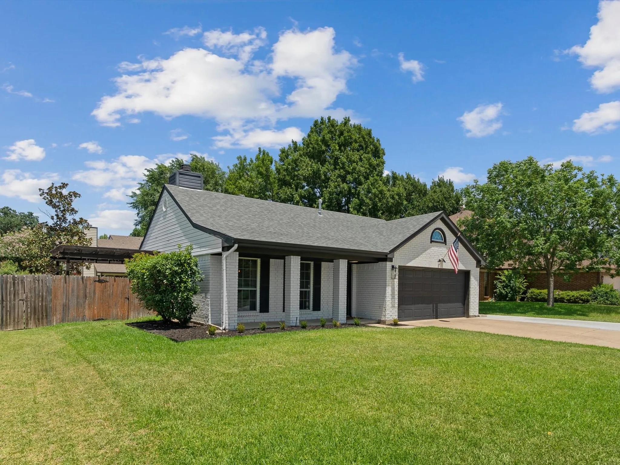 Single-story residence featuring light-colored brick and siding, a dark-toned shingle roof, a prominent attached garage with a dark door, and a well-maintained front lawn with established landscaping