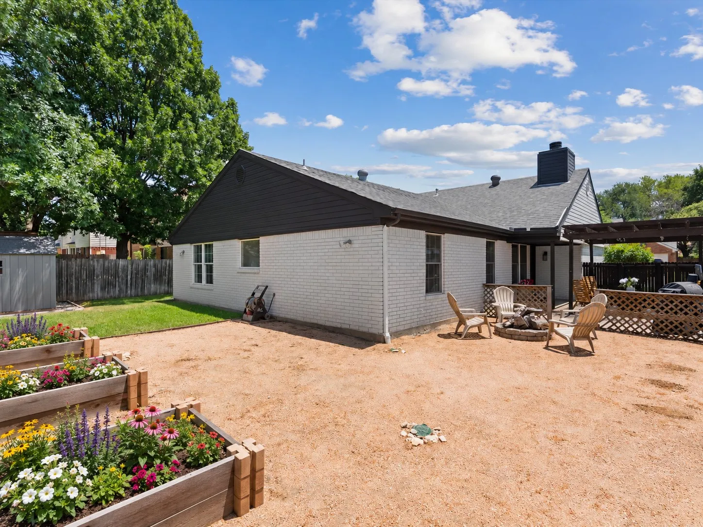 White brick exterior with black trim, gravel patio area, wood deck with pergola, raised garden beds, and a manicured lawn