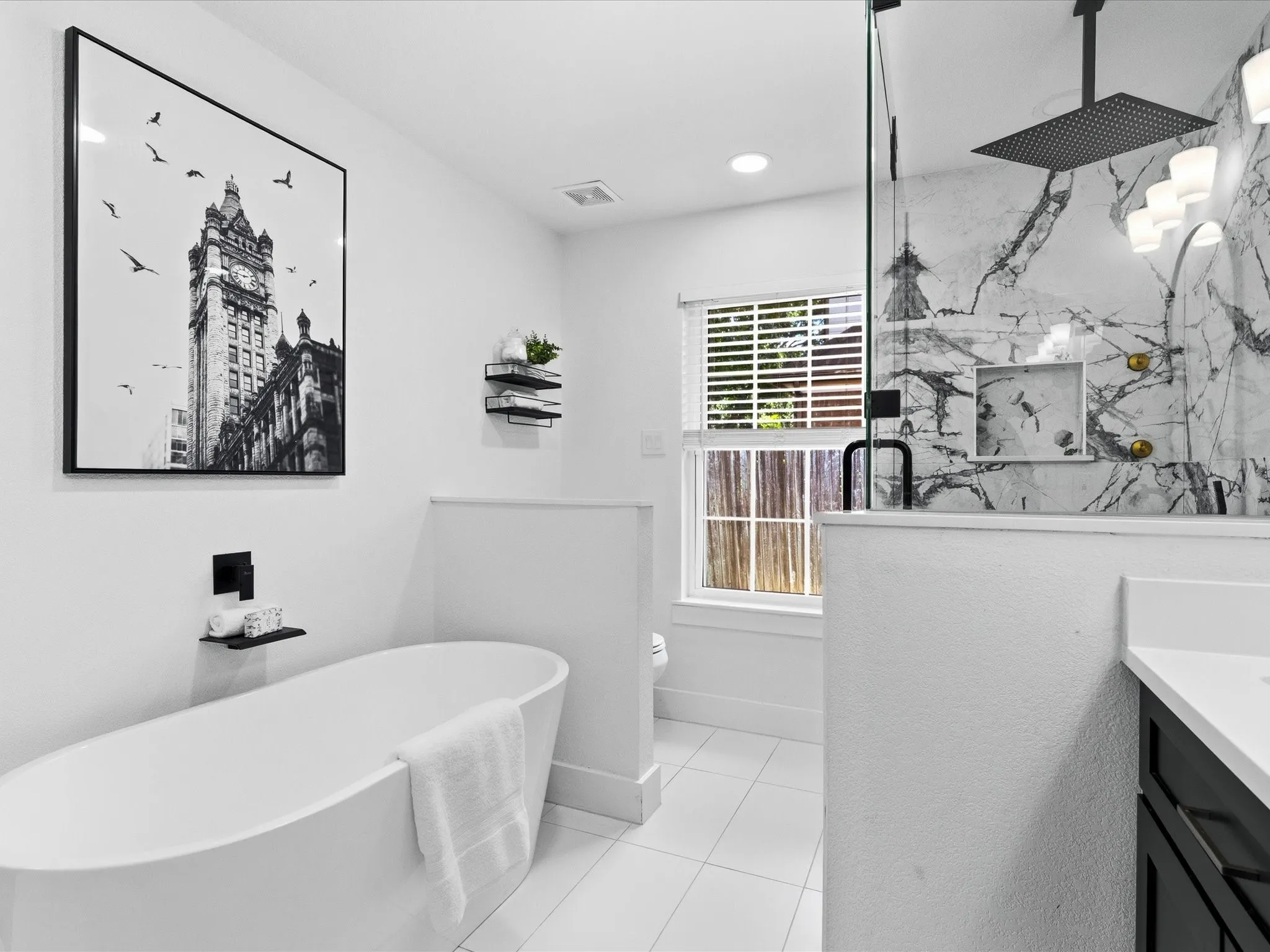 Freestanding soaking tub, walk-in shower with glass enclosure, large format wall tile with visible veining, matte black shower fixtures, and white tile flooring
