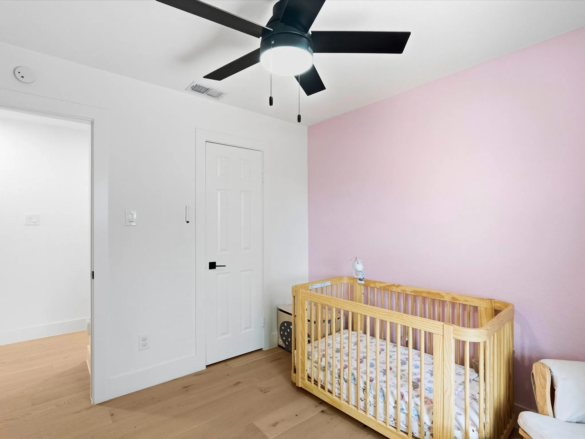 Bright nursery featuring soft pink accent wall, natural wood crib, ceiling fan with integrated lighting, white six-panel doors, and warm wood-finish flooring throughout