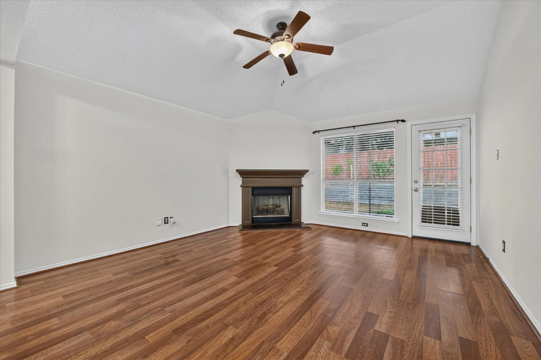 Spacious living room showcasing warm wood-look flooring, neutral wall paint, wood burning fireplace and a ceiling fan with lights creating an inviting space for relaxation.