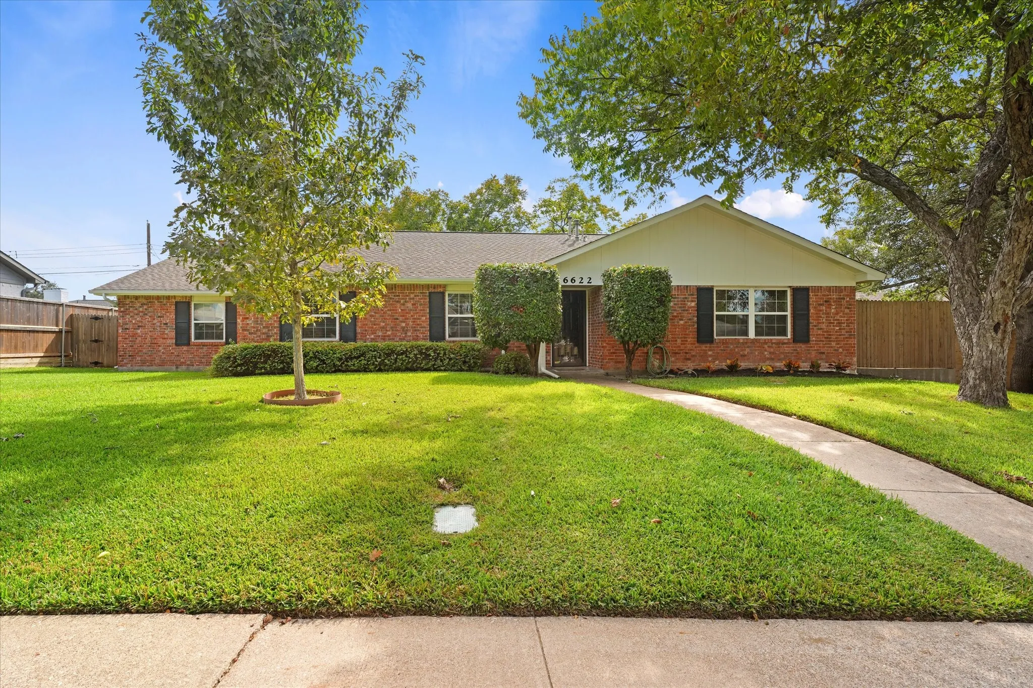Single-story brick exterior featuring a low-pitch roof, symmetrical window placement with dark shutters, and a front lawn with mature trees