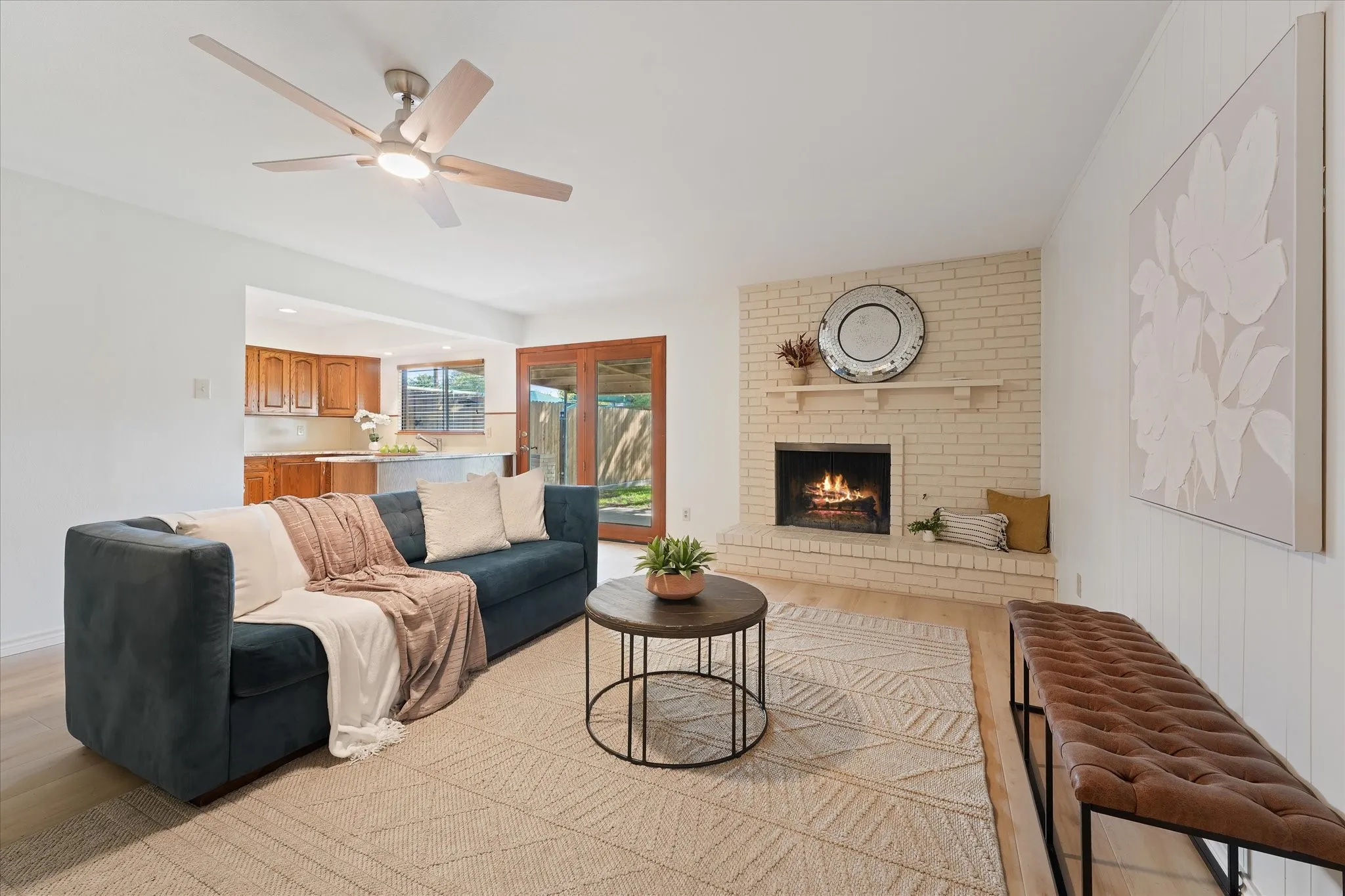 Living area featuring a floor-to-ceiling brick fireplace with a raised hearth, wood-finish LVP flooring, a multi-blade ceiling fan with integrated lighting, and glass-paneled patio doors.  Ready for a cool fall evening in front of the fireplace.