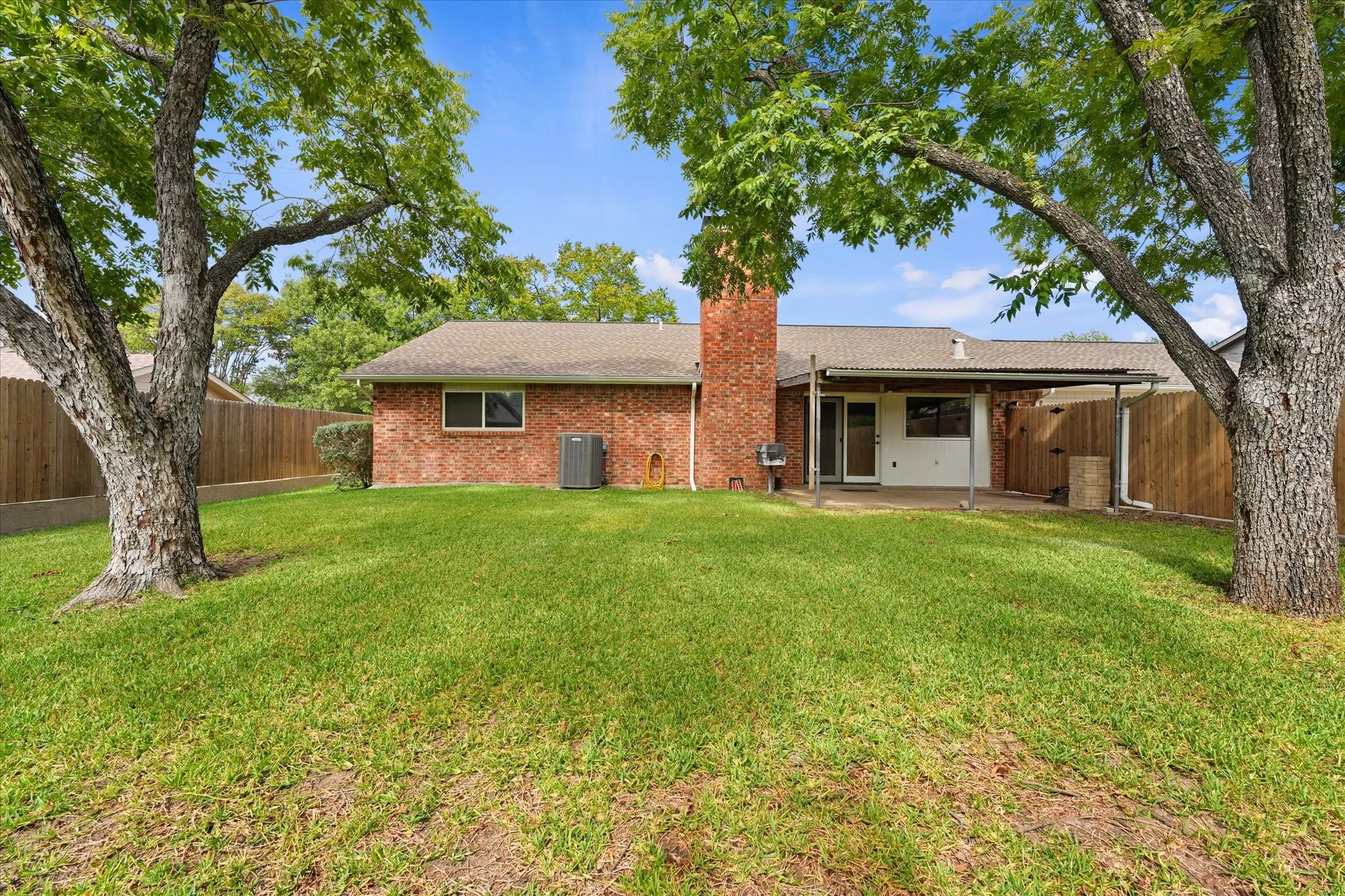 Exterior featuring brick siding, a covered patio, a brick chimney, and mature trees situated on a lawn enclosed by wood fencing