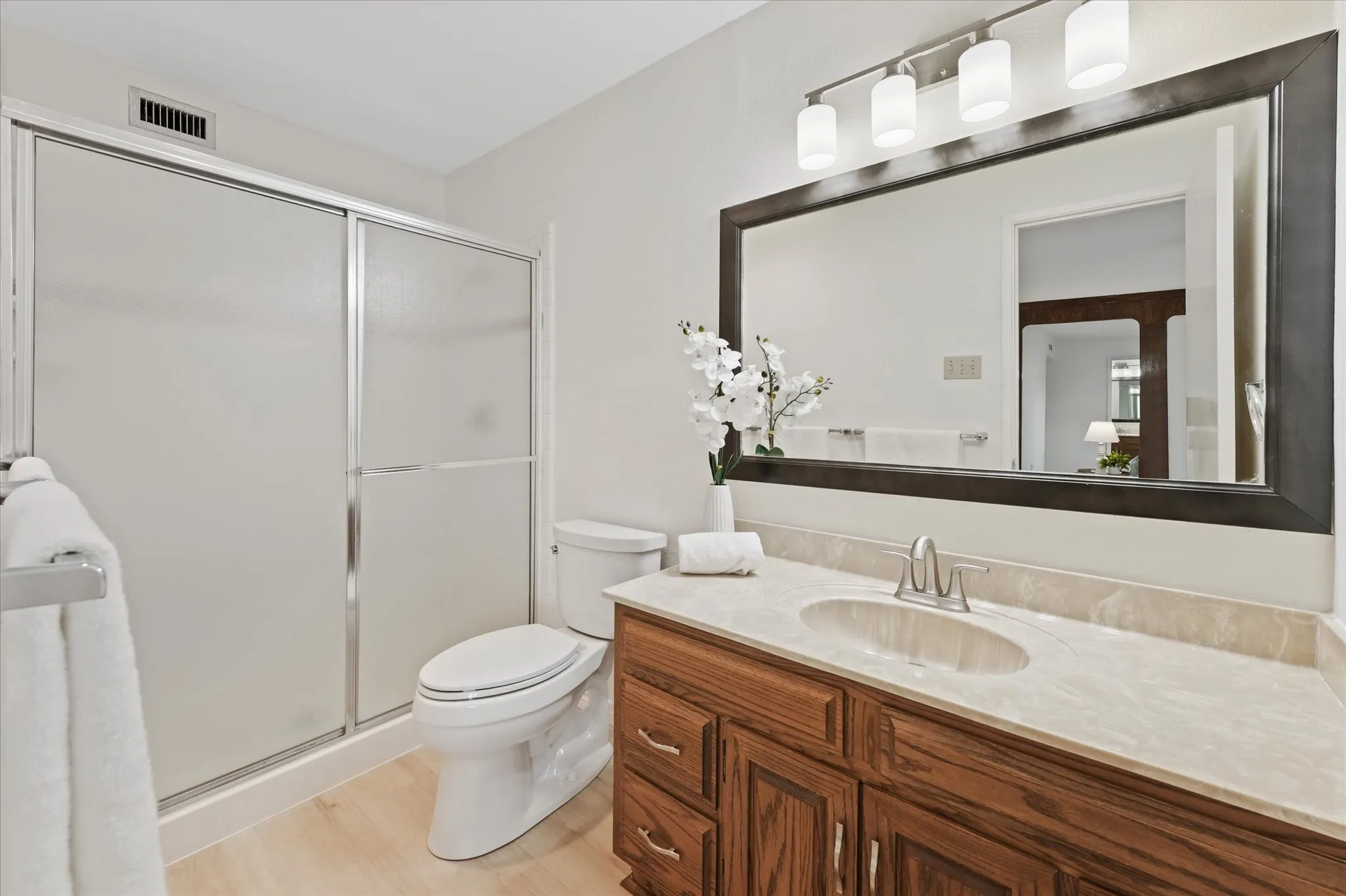 Bathroom featuring a wooden vanity with oval undermount sink, updated faucet and cabinet hardware a framed wall mirror, new four-light vanity fixture, and a shower with sliding glass doors
