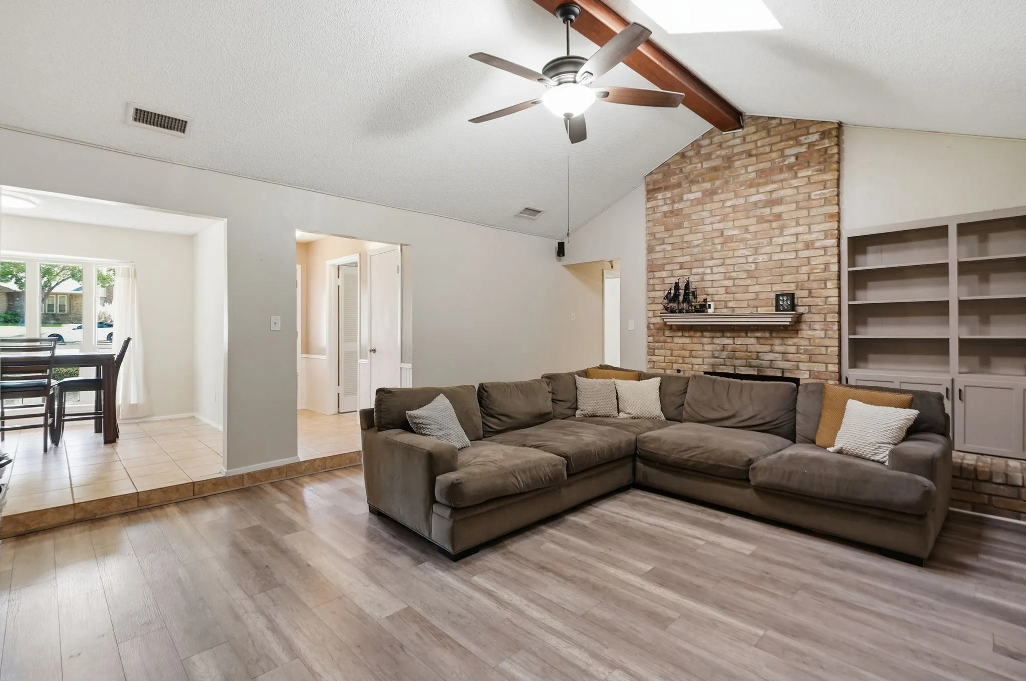 Vaulted ceiling with exposed beam, wood-finish flooring, brick accent wall with mantel, ceiling fan, and built-in shelving