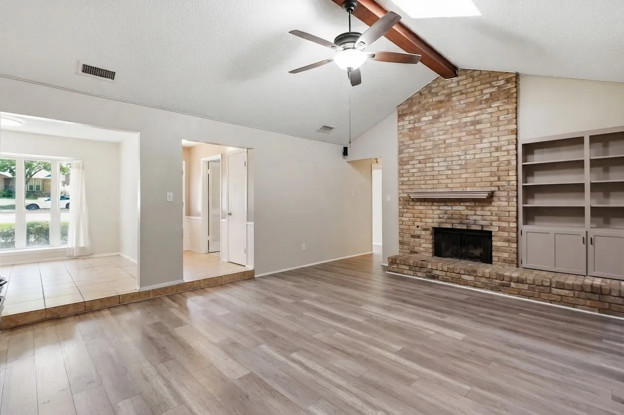 Vaulted ceiling with wood beam, wood-finish flooring, brick fireplace with mantel, built-in shelving, and a ceiling fan