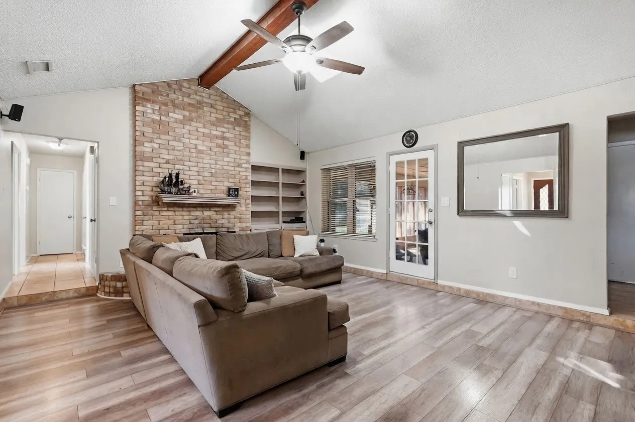 Vaulted ceiling with exposed wood beam, wood-finish flooring, brick-clad fireplace with mantel, built-in shelving, and a ceiling fan with integrated lighting