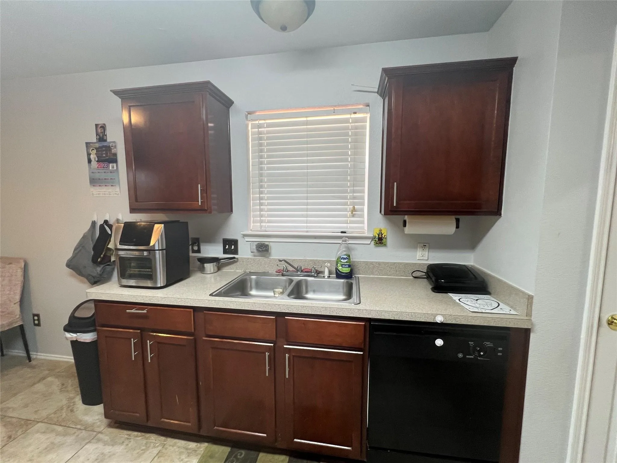 Wood-finish cabinetry with brushed nickel hardware, light-toned countertop, double basin stainless steel sink, and a black built-in dishwasher