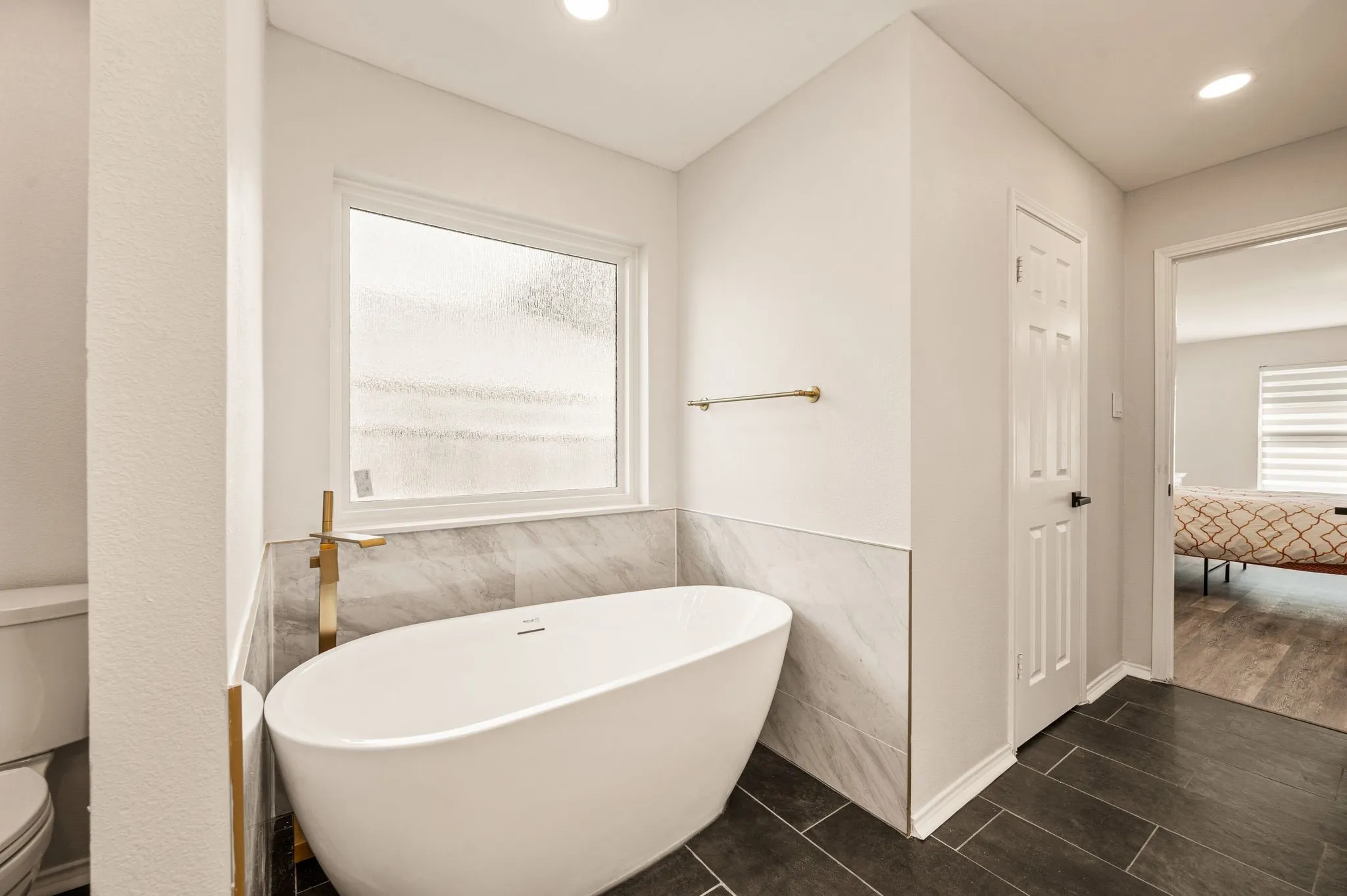 Freestanding soaking tub with a floor-mounted gold-tone faucet, complemented by large format tile flooring and a privacy window