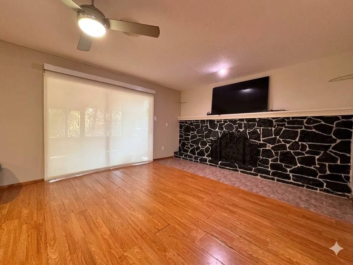 Wood-finish flooring, stone-clad fireplace with metal grate, ceiling fan with integrated lighting, and window with roller shade