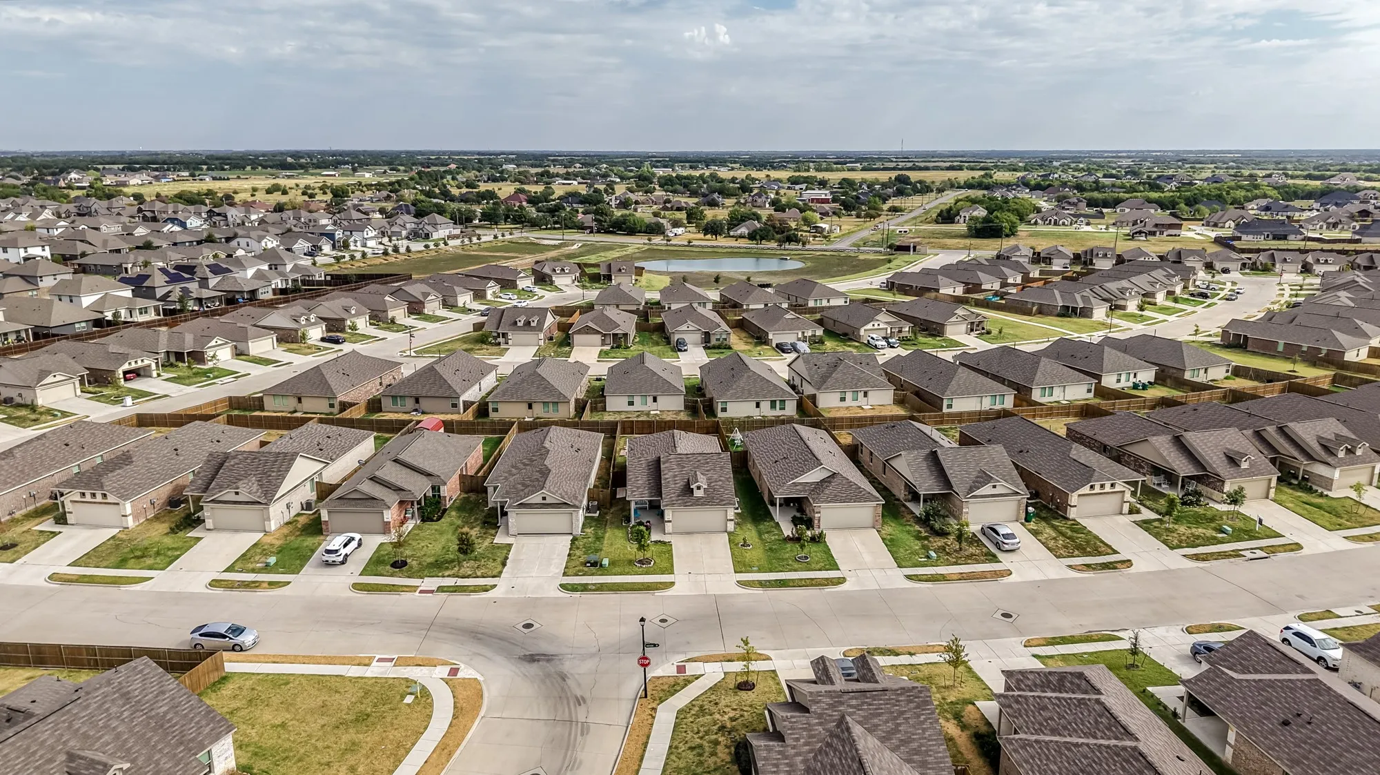 Aerial perspective showcasing a residential community with multiple single-family homes, paved streets, and a central pond feature