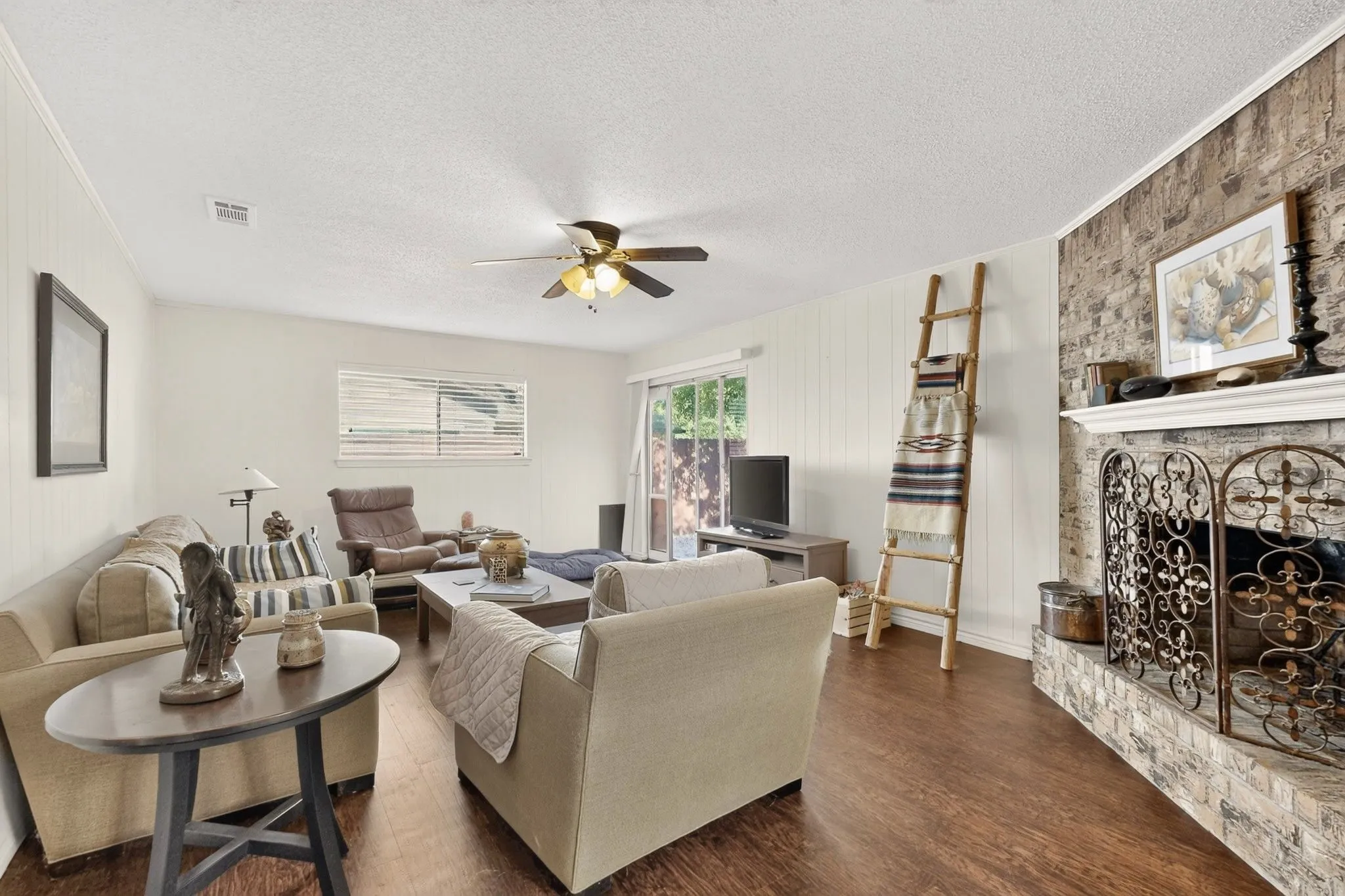 Living area featuring wood-finish flooring, a ceiling fan, a brick-front fireplace with a decorative screen, and vertical paneling