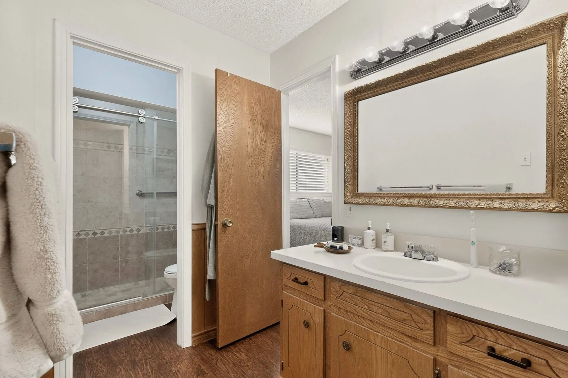 Wood-finish flooring, vanity with white countertop and integrated sink, decorative framed mirror, and a shower with sliding glass door and tiled surround