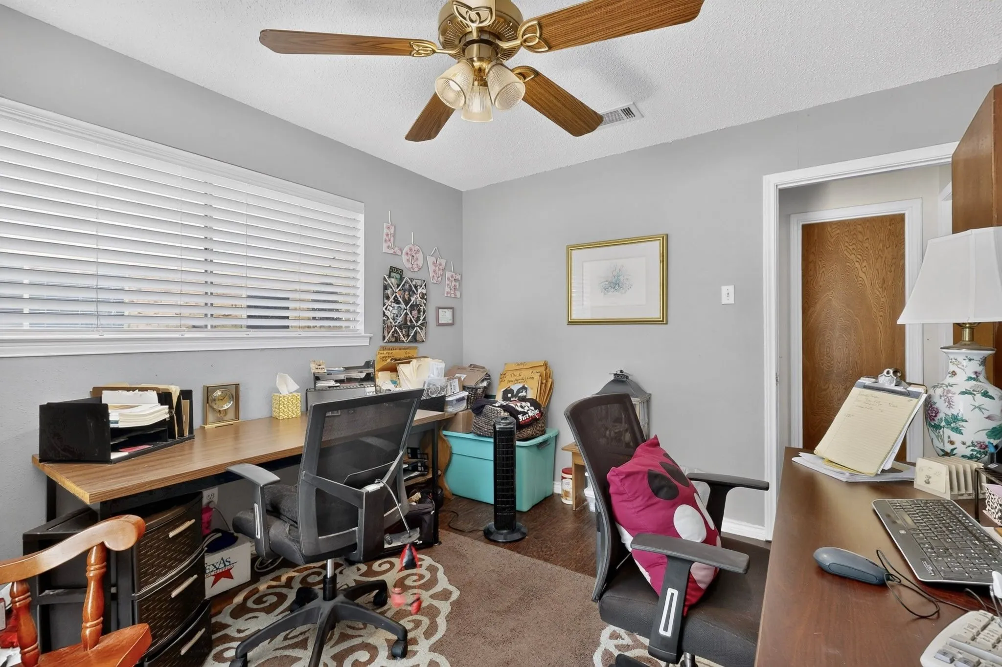 Recessed window with horizontal blinds, overhead ceiling fan with wood-finish blades, light gray painted walls, and a doorway with a wood-finish door