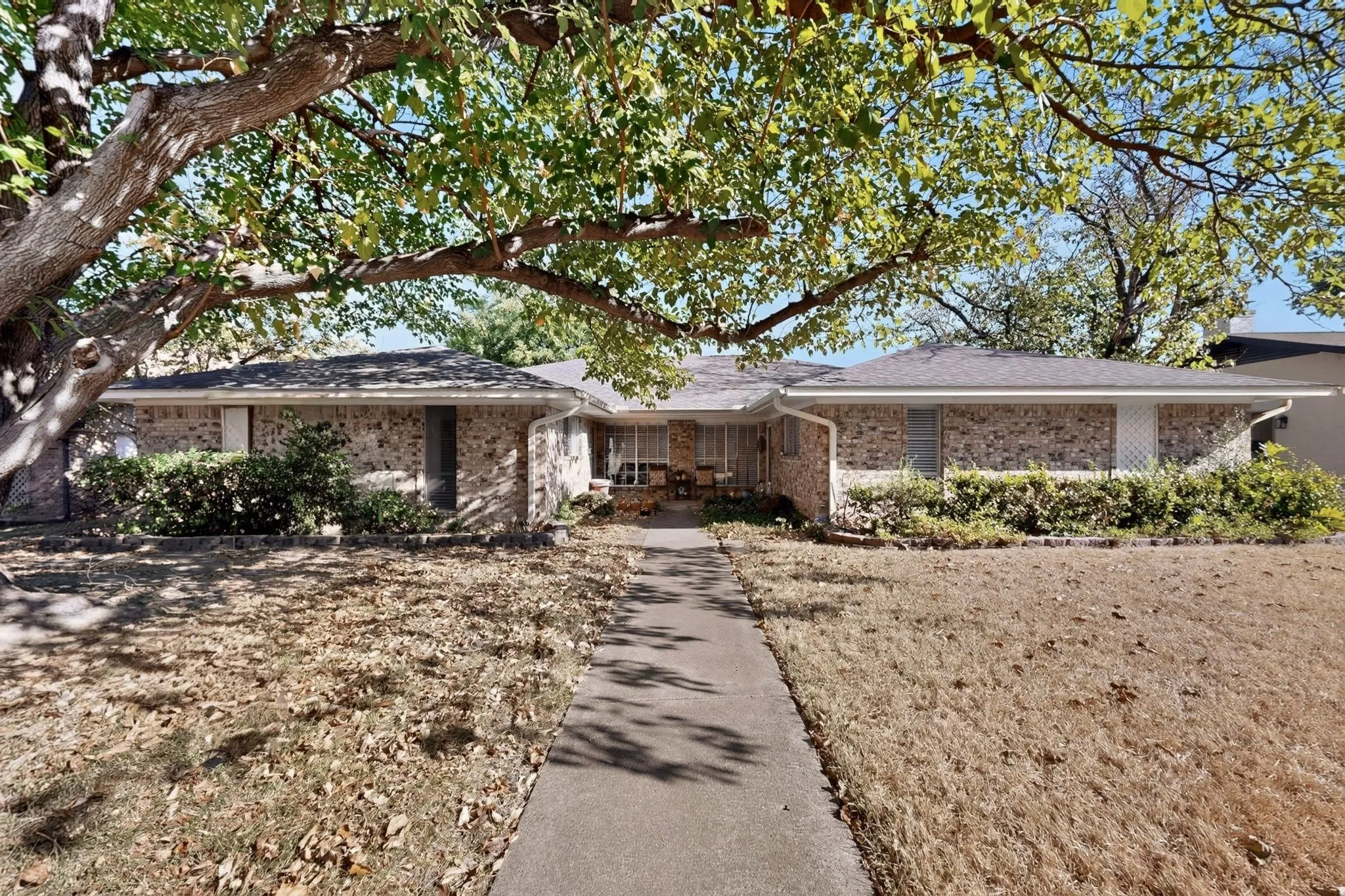 Brick exterior residence with a gray shingle roof, mature trees, and a concrete walkway