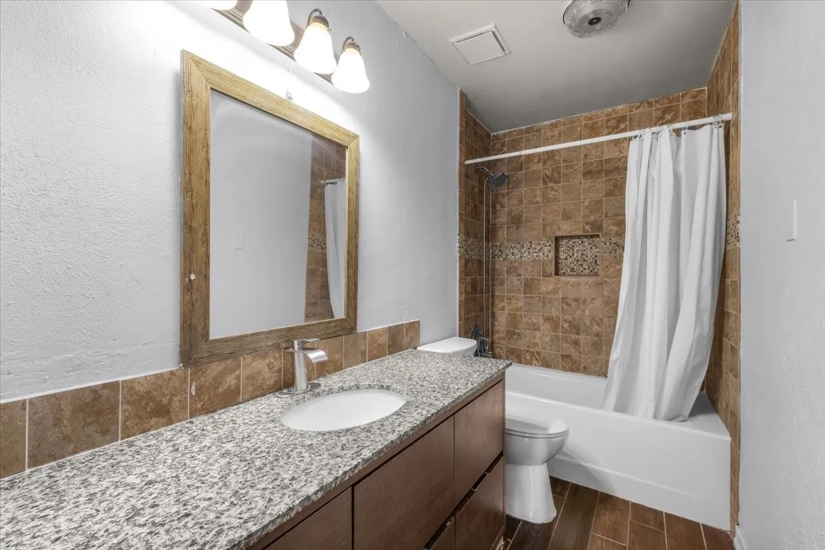 Bathroom featuring a granite-style countertop, wood-finish vanity, and tiled shower surround with a decorative accent band