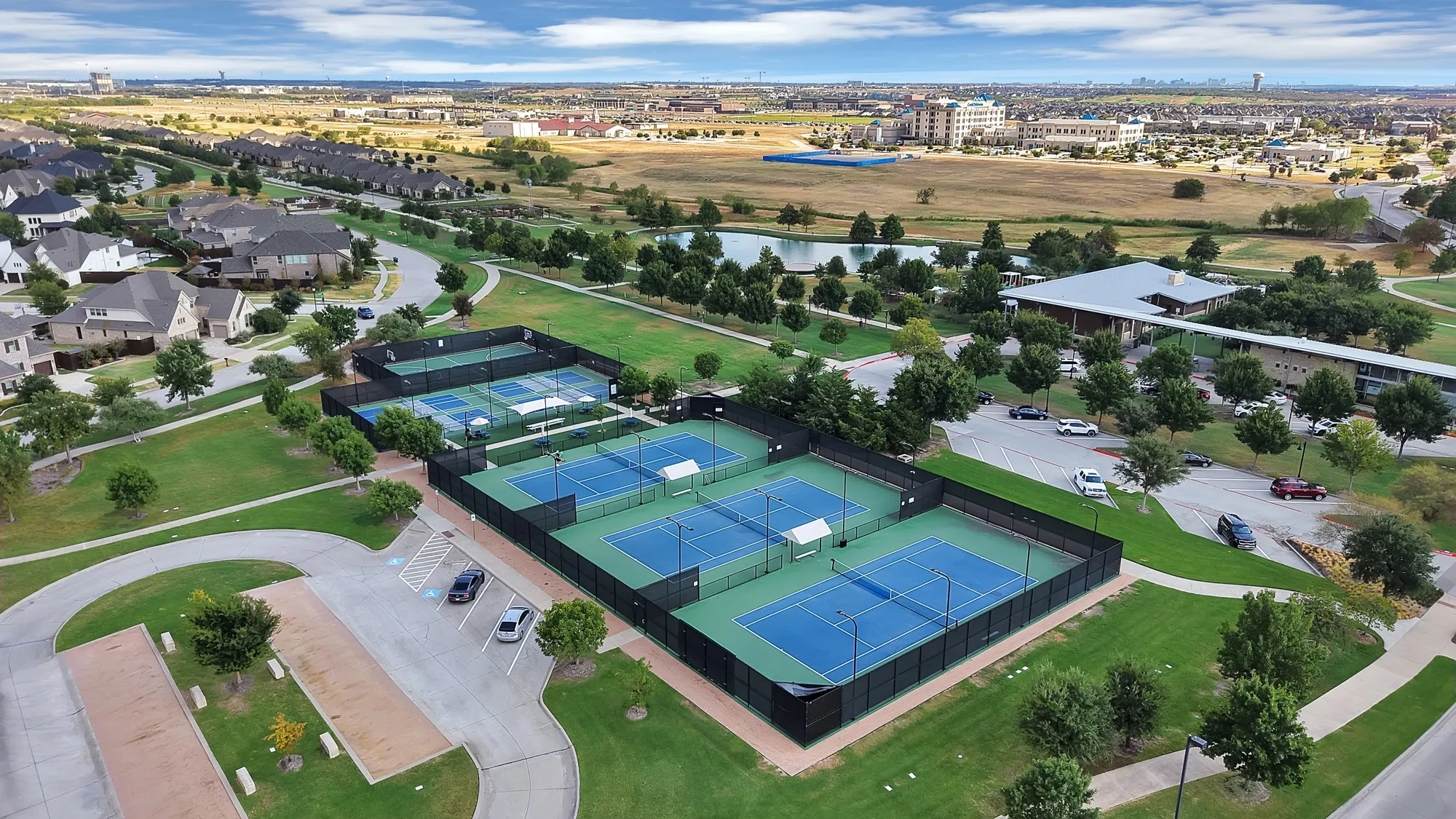 Expansive recreational area featuring multiple tennis courts, a large community building with an extended roofline, a tranquil pond, and surrounding green spaces