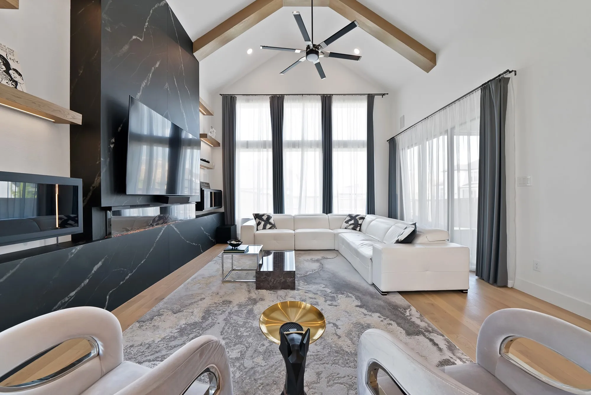 Vaulted ceiling with exposed wood beams, large format black stone accent wall, ceiling fan with integrated lighting, wood-finish flooring, and expansive windows with vertical blinds