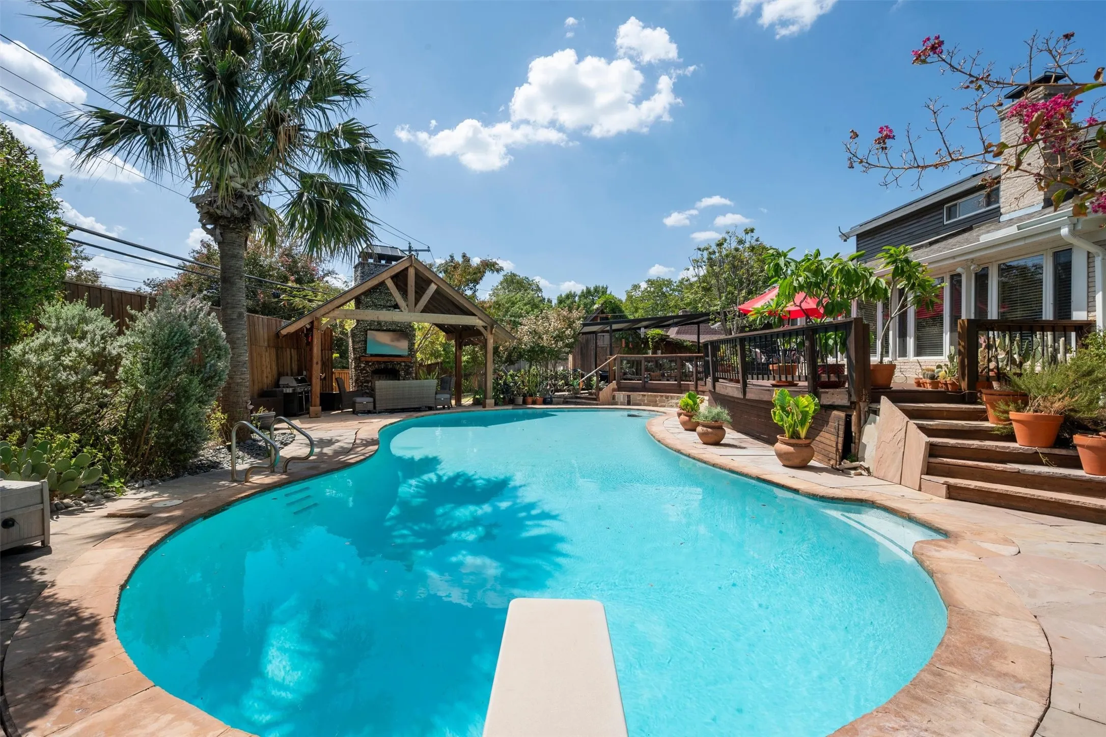 Curved swimming pool with a diving board, adjacent stone paver decking, a wood-framed outdoor living structure with a built-in fireplace, and a multi-level wood deck with​​‌​​​​‌​​‌‌​‌‌​​​‌‌​‌​‌​‌​​​‌​​ steps