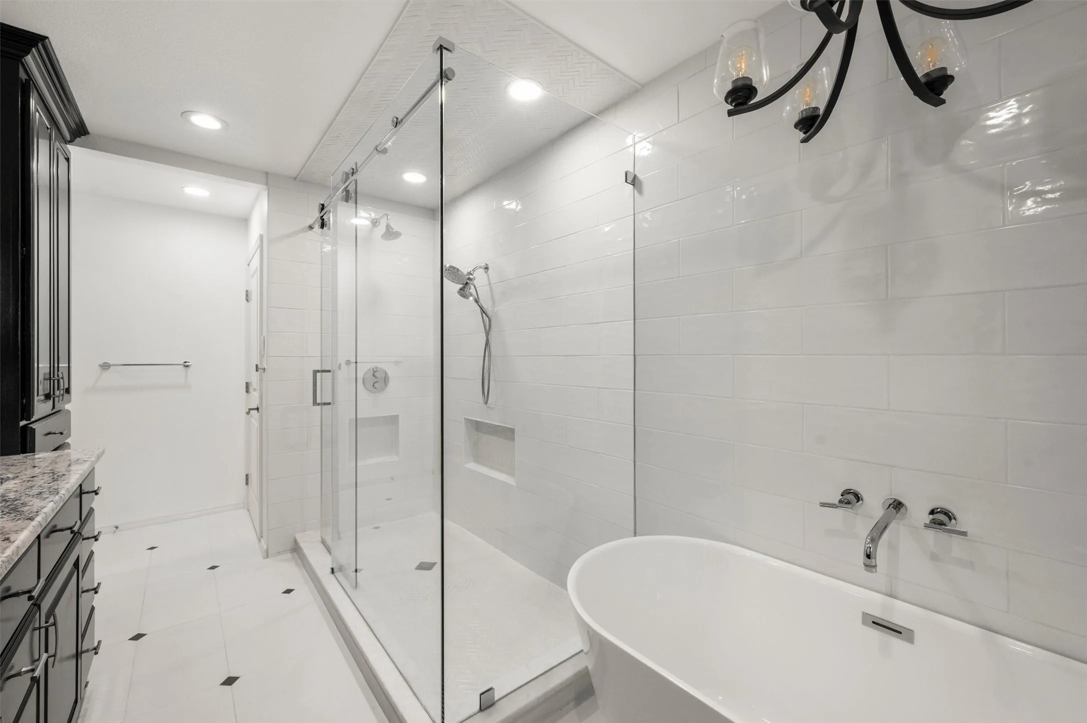 Freestanding soaking tub, frameless glass shower enclosure with dual showerheads, dark wood vanity with stone countertop, recessed lighting, and a decorative chandelier