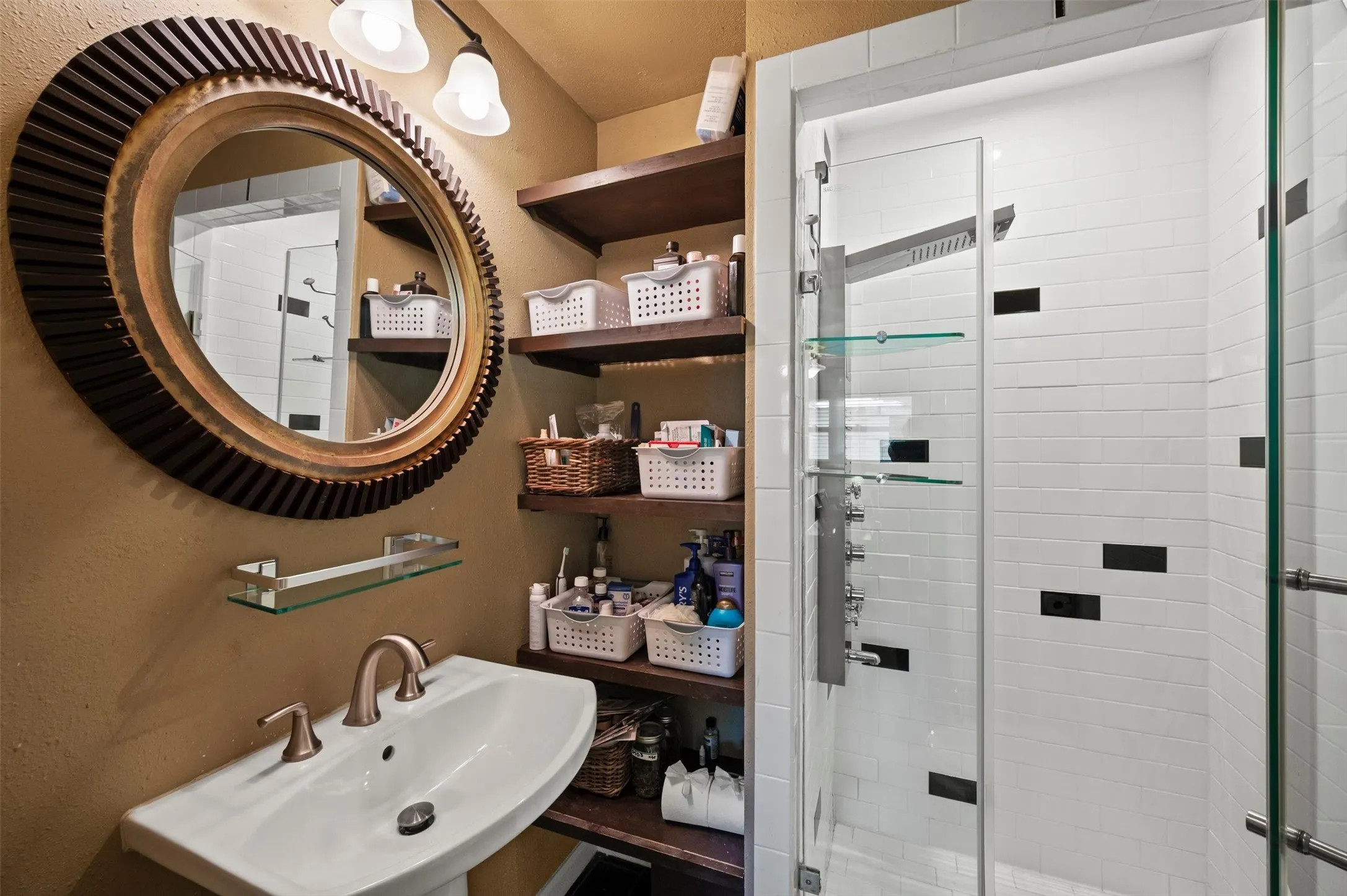 Pedestal sink with brushed metal faucet, open wood-finish shelving, and a glass-enclosed shower with white tiled walls