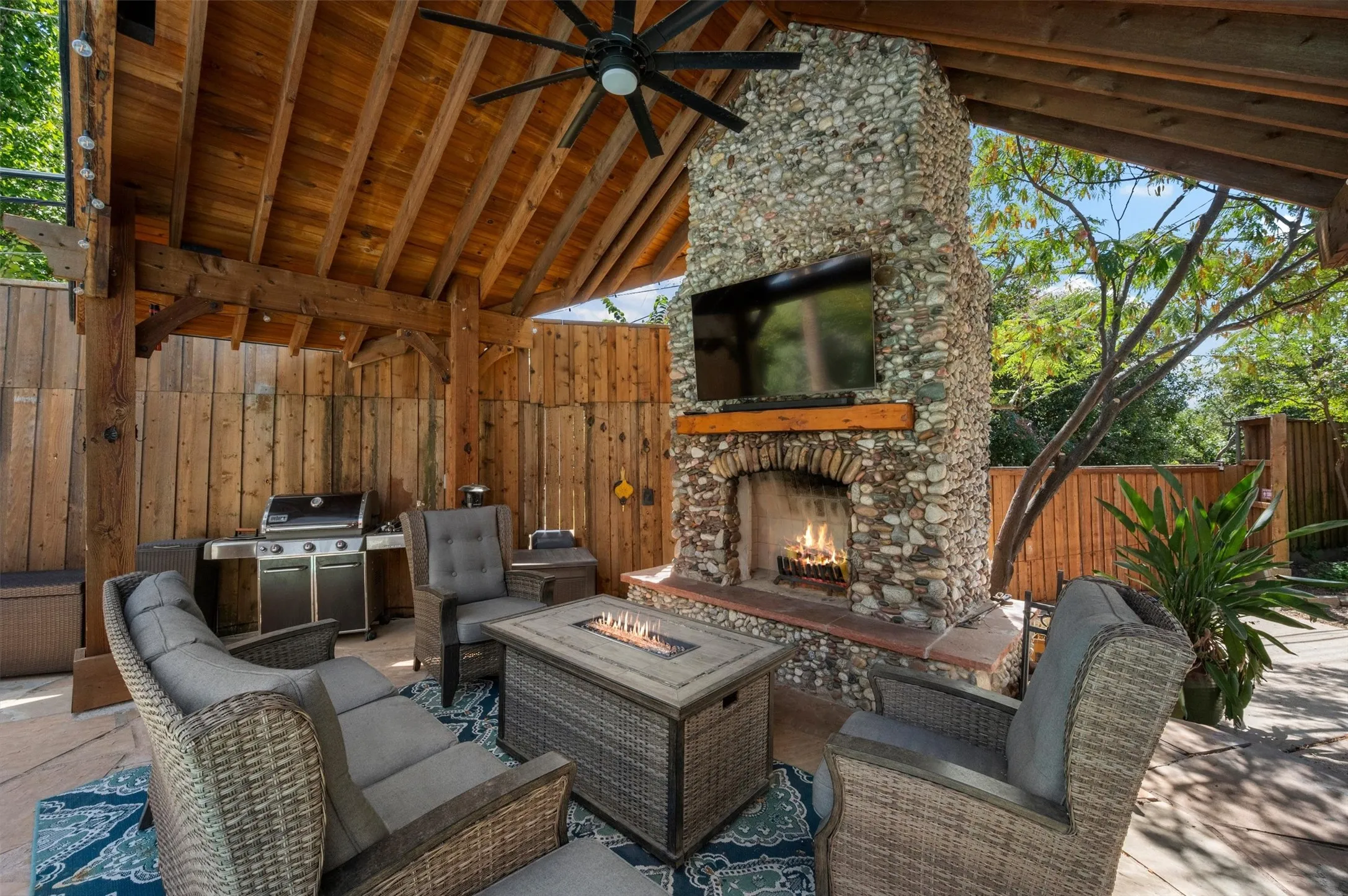 Covered outdoor patio featuring a stone fireplace, wood-beamed ceiling, ceiling fan, and built-in grill