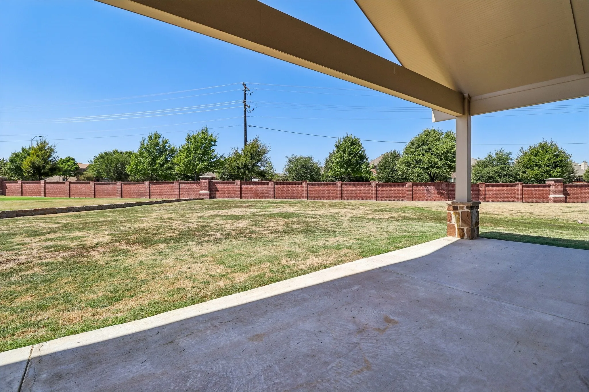 Covered concrete patio with stone-clad pillar and expansive lawn, bordered by a brick privacy wall
