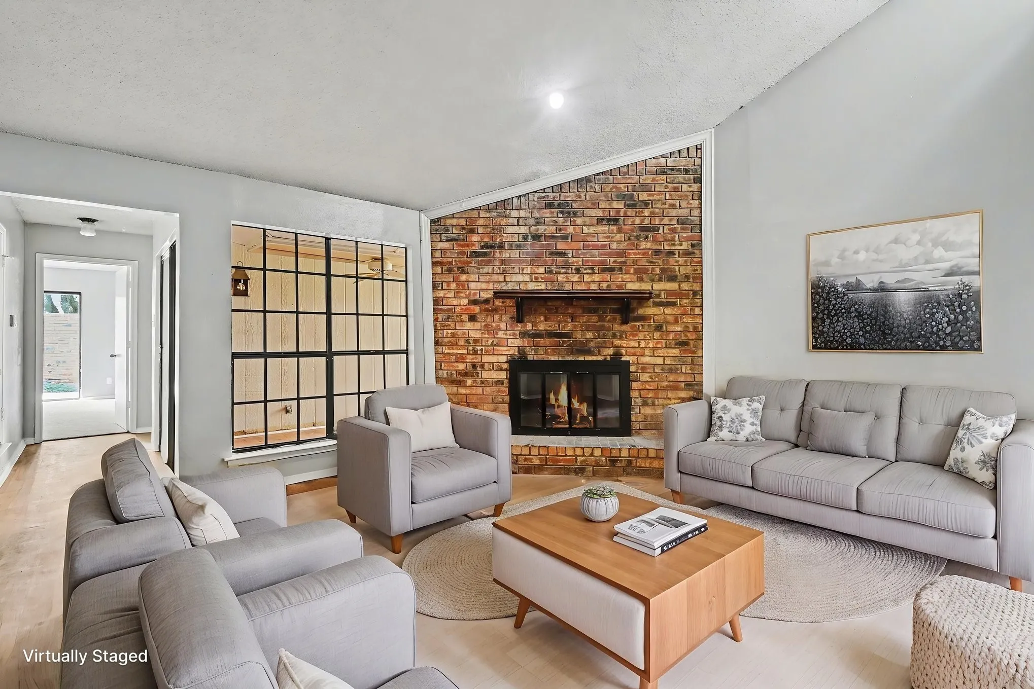 Brick fireplace with hearth, pitched ceiling, large grid-pattern window, and wood-finish flooring