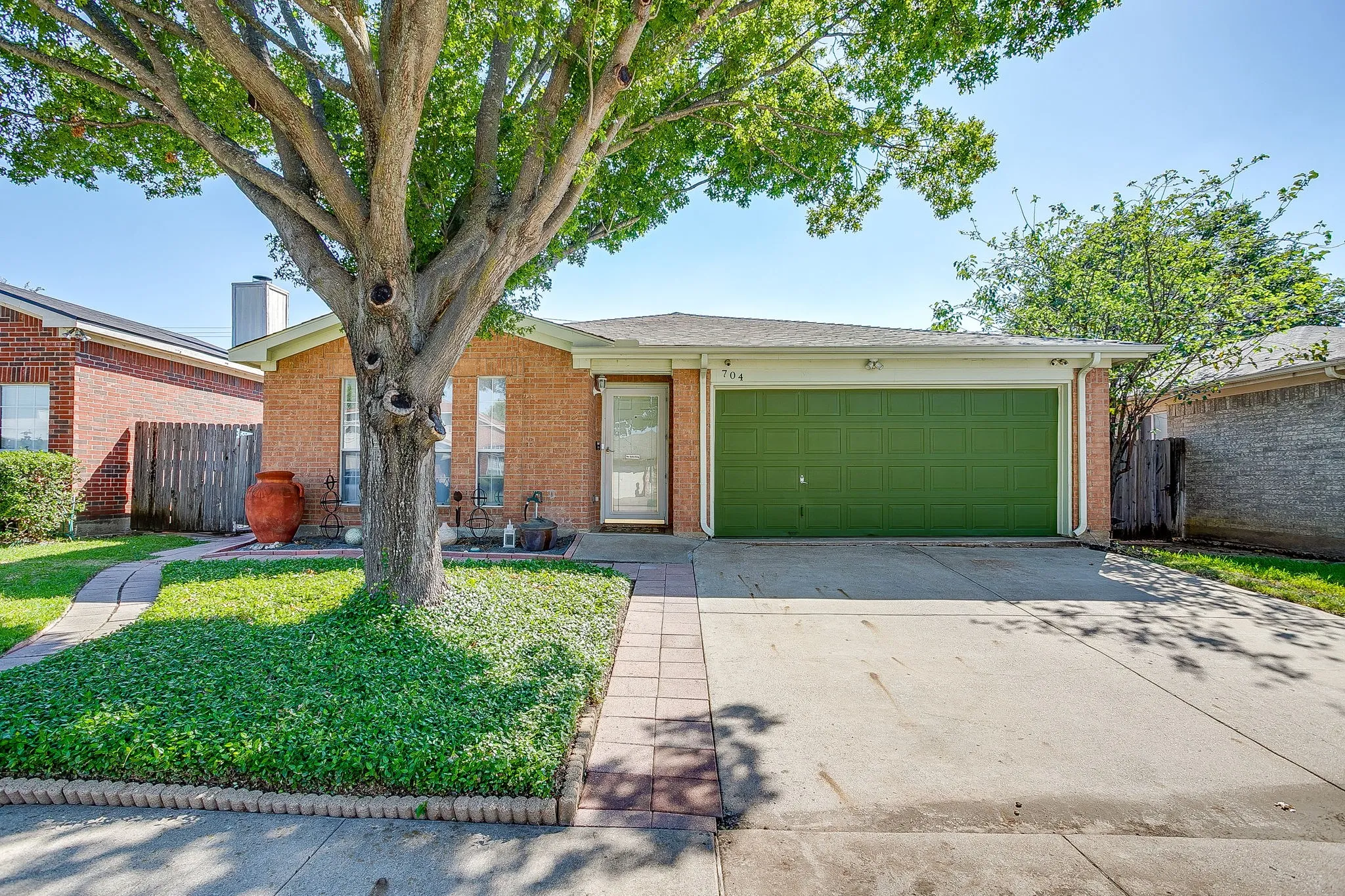 Brick exterior facade, two-car garage with solid door, concrete driveway, front yard featuring mature deciduous tree, and paved stone walkway