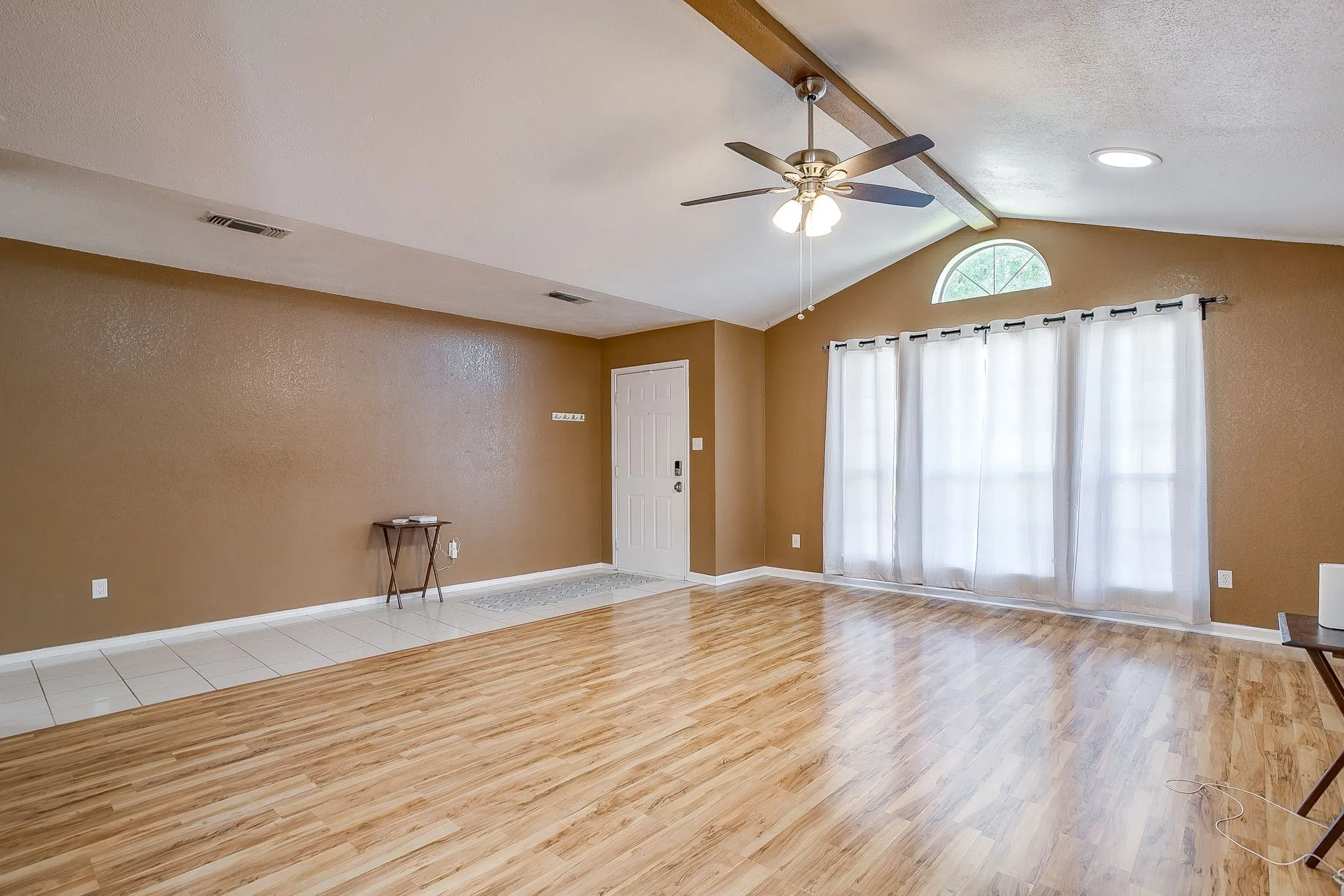 Vaulted ceilings featuring an exposed beam, wood-finish flooring, a multi-pane window with an arched transom, and a central ceiling fan