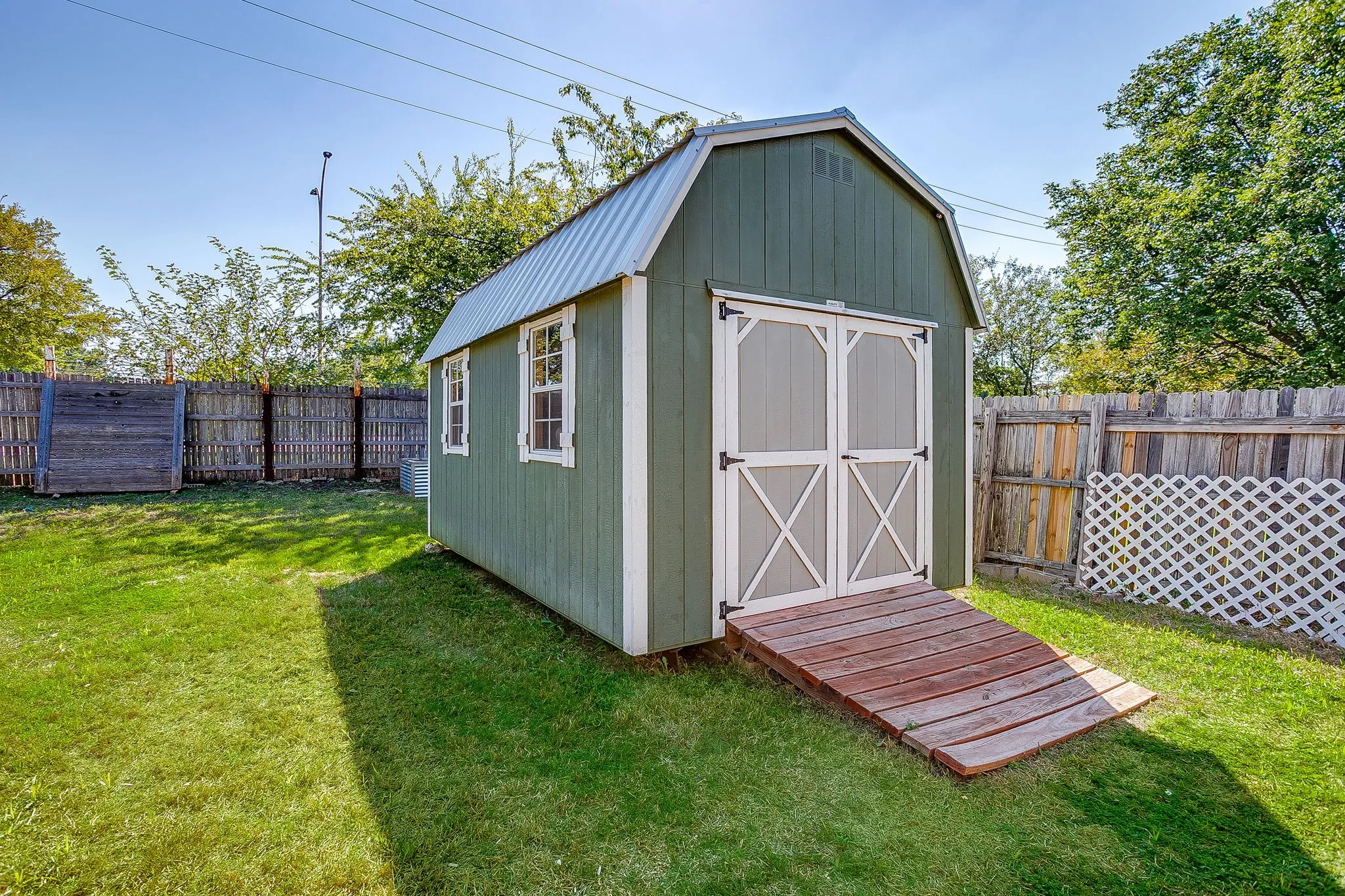 Storage structure featuring a gambrel roof, double-door entry, wood-plank ramp, and exterior windows, set within a fenced yard