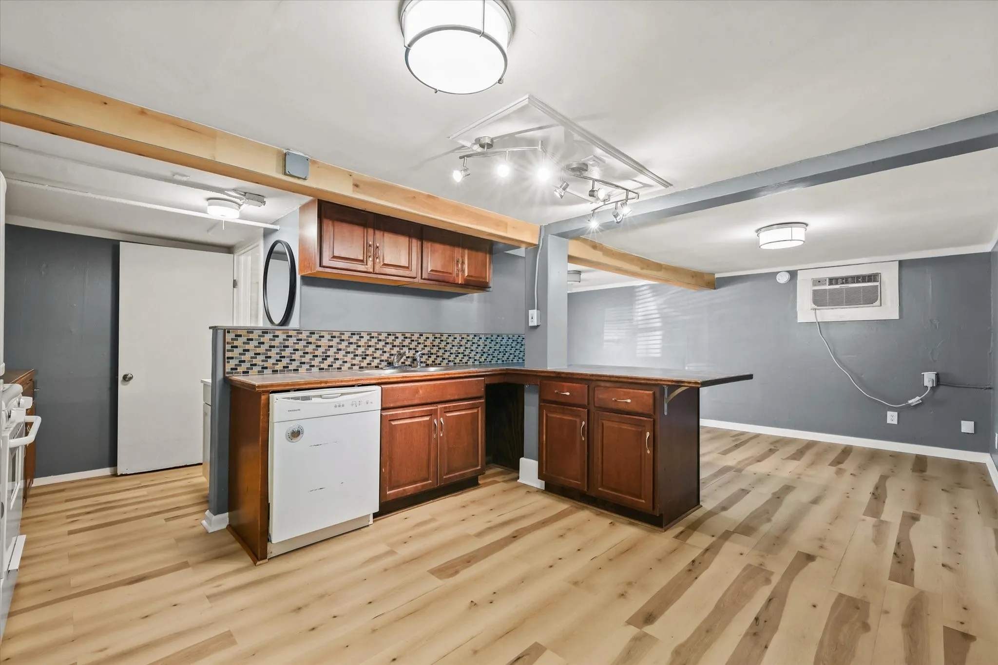Wood-finish flooring, wood cabinetry, patterned tile backsplash, track lighting, and a white dishwasher