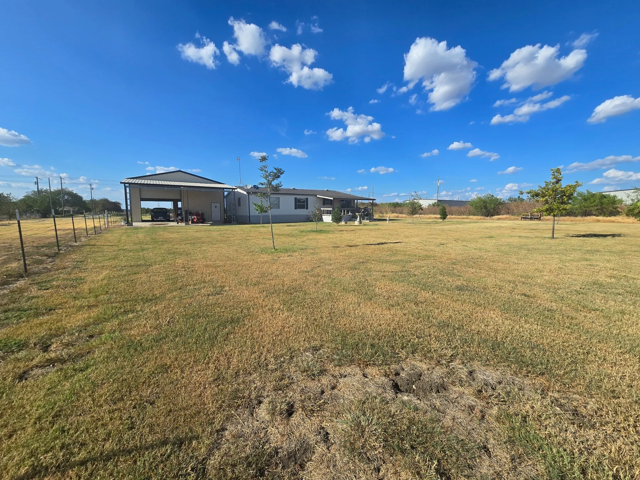 Expansive backyard outdoor area featuring a metal-framed carport, a single-story residence, and a wire fence bordering the property