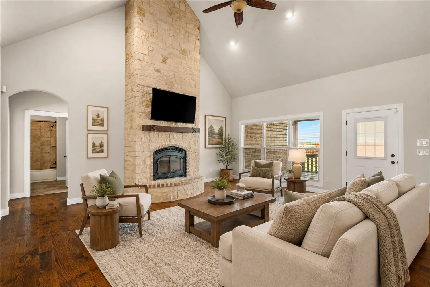 Vaulted ceiling, stone fireplace with hearth, wood-finish flooring, ceiling fan, and transom windows
