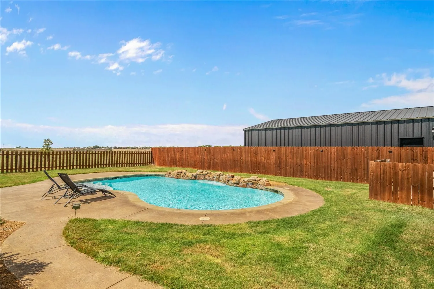 Curved swimming pool with rock detailing, surrounded by a concrete patio, and a grass lawn with wood fencing