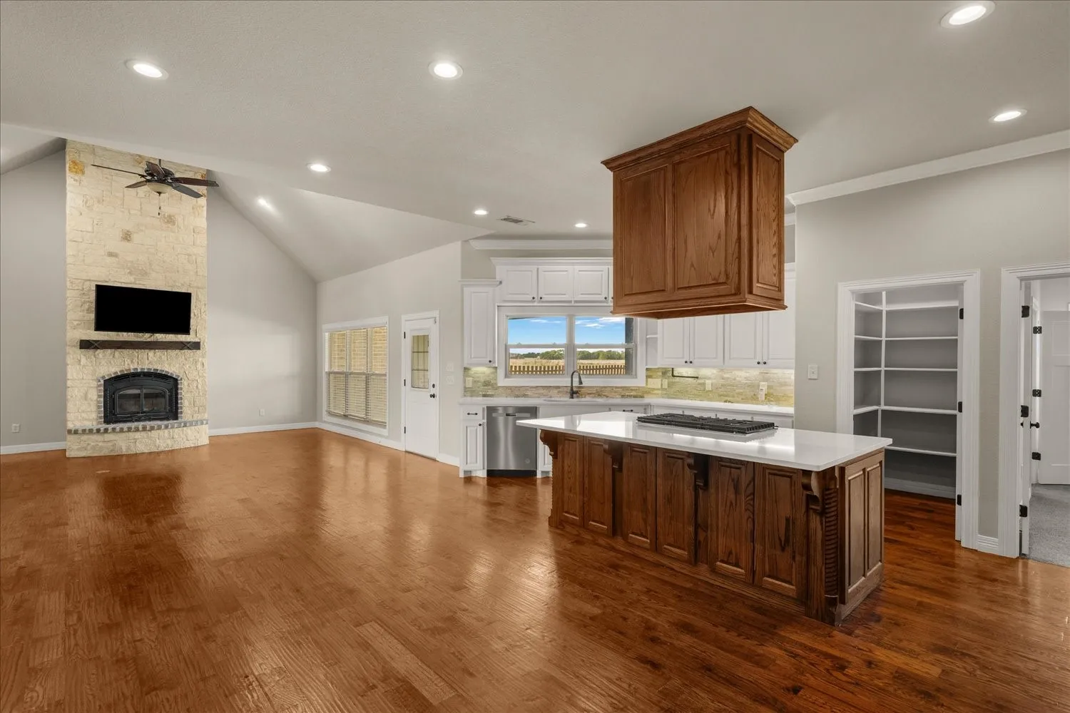 Vaulted ceiling, stone fireplace with a wood-beam mantel, wood-finish flooring, kitchen island with built-in cooktop, and white cabinetry