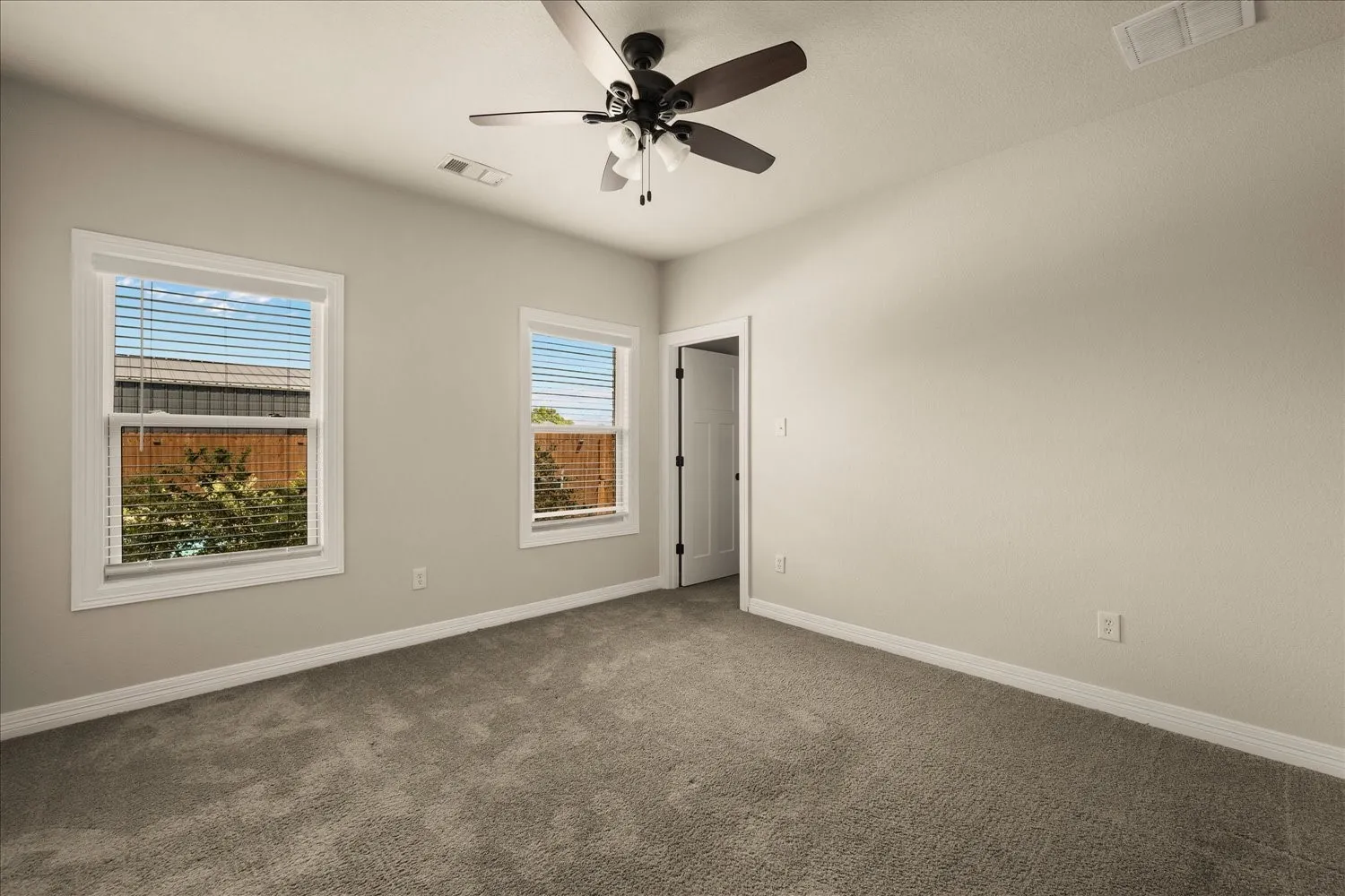 Spacious room featuring neutral carpeting, two double-hung windows with blinds, a ceiling fan with integrated lighting, and a white interior door