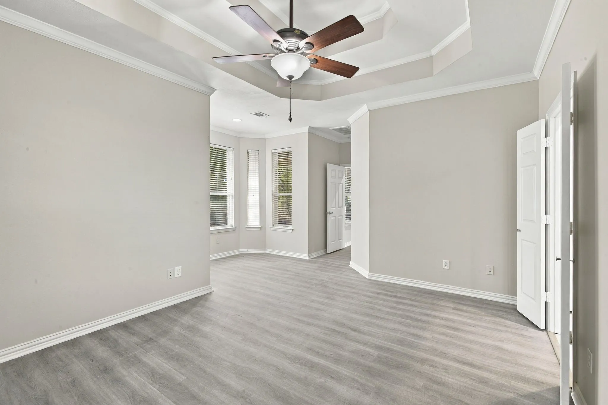 Tray ceiling with crown molding, wood-finish flooring, ceiling fan, bay windows, and white trim