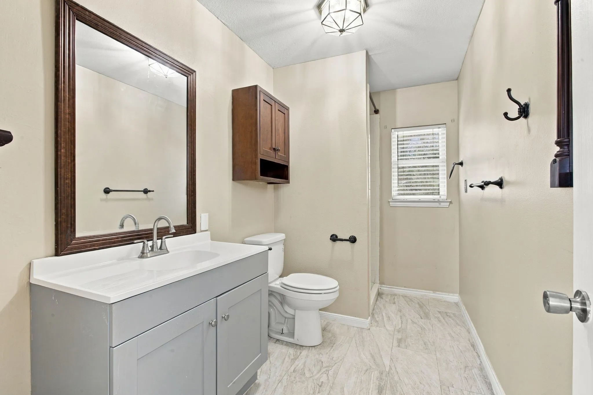 Single vanity with a white countertop and gray cabinetry, wood-finish framed mirror, wood-finish wall-mounted storage cabinet, and light-toned tile flooring