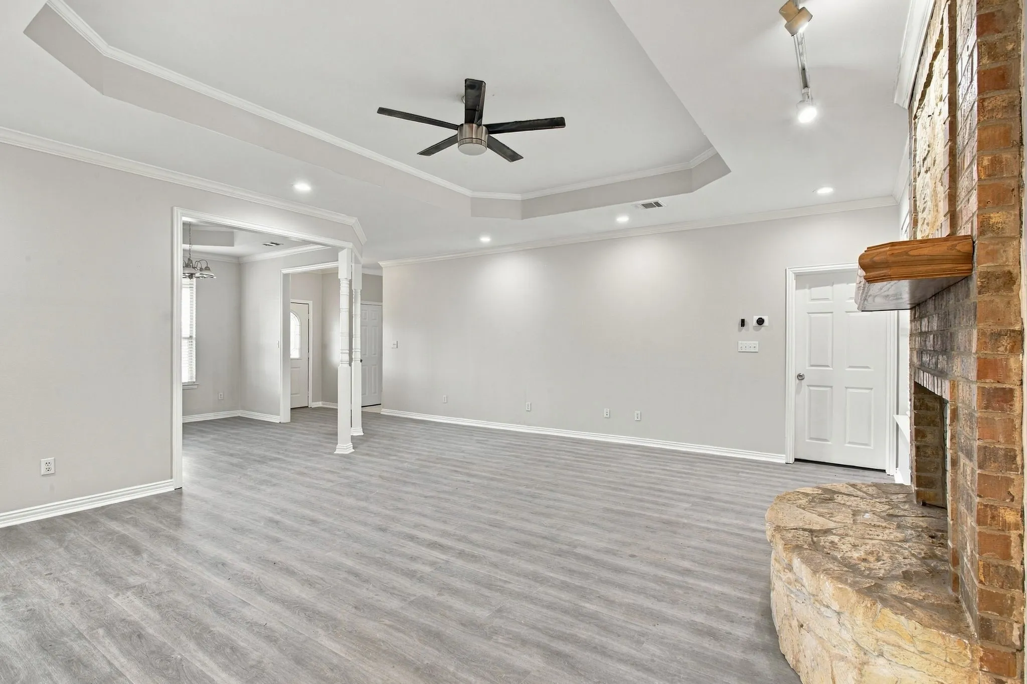 Spacious room featuring wood-finish flooring, a tray ceiling with a ceiling fan, recessed lighting, and an exposed brick fireplace with a stone hearth