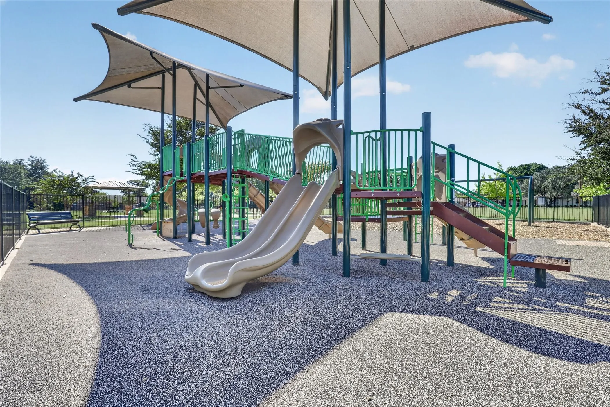 Playground featuring a tan shade structure, multi-level play equipment with slides, a soft-surface ground cover, and surrounding black fencing