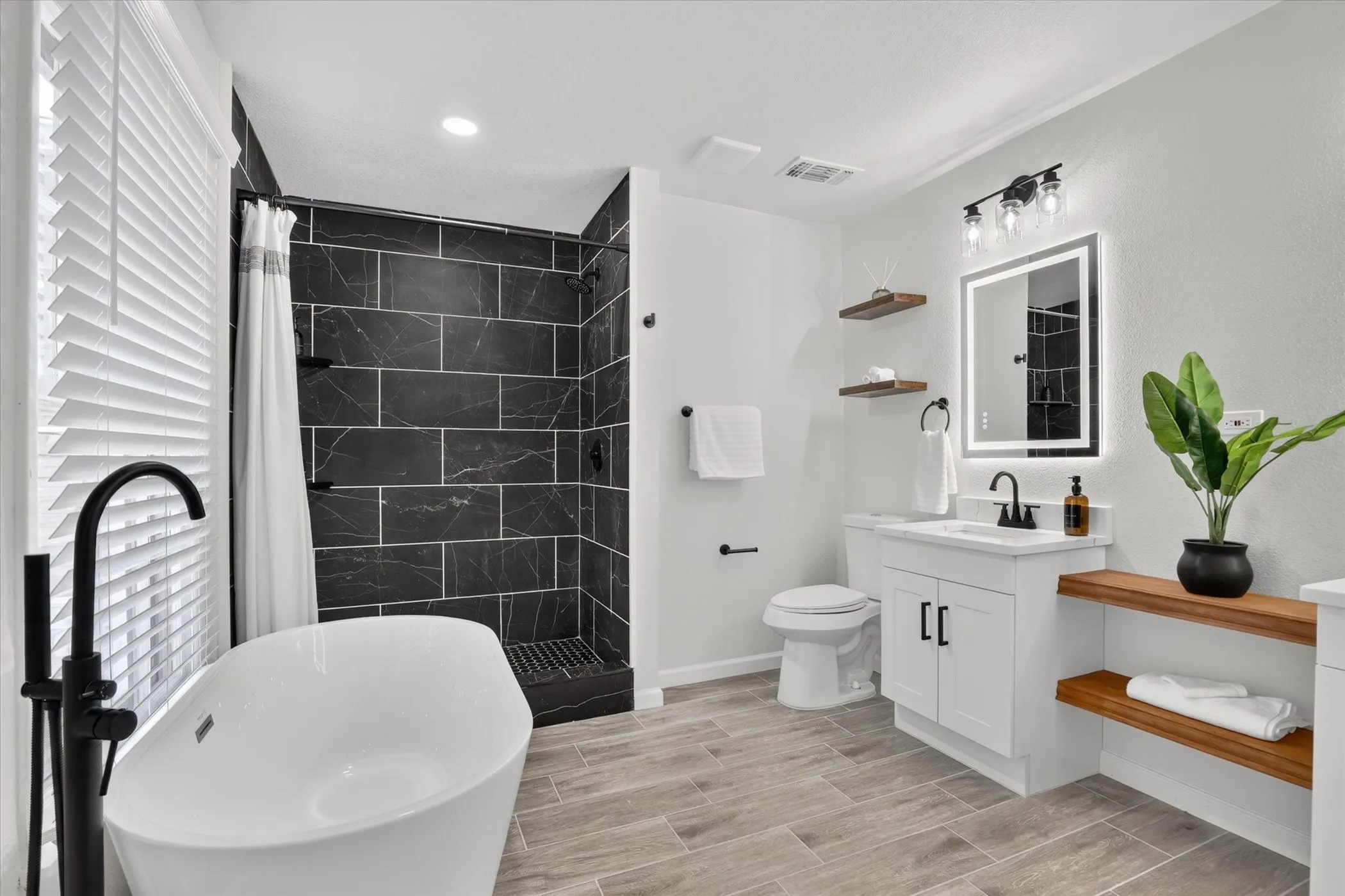 Freestanding soaking tub with a floor-mounted matte black faucet, walk-in shower with dark subway tile, single vanity with a white countertop, and wood-finish tile flooring