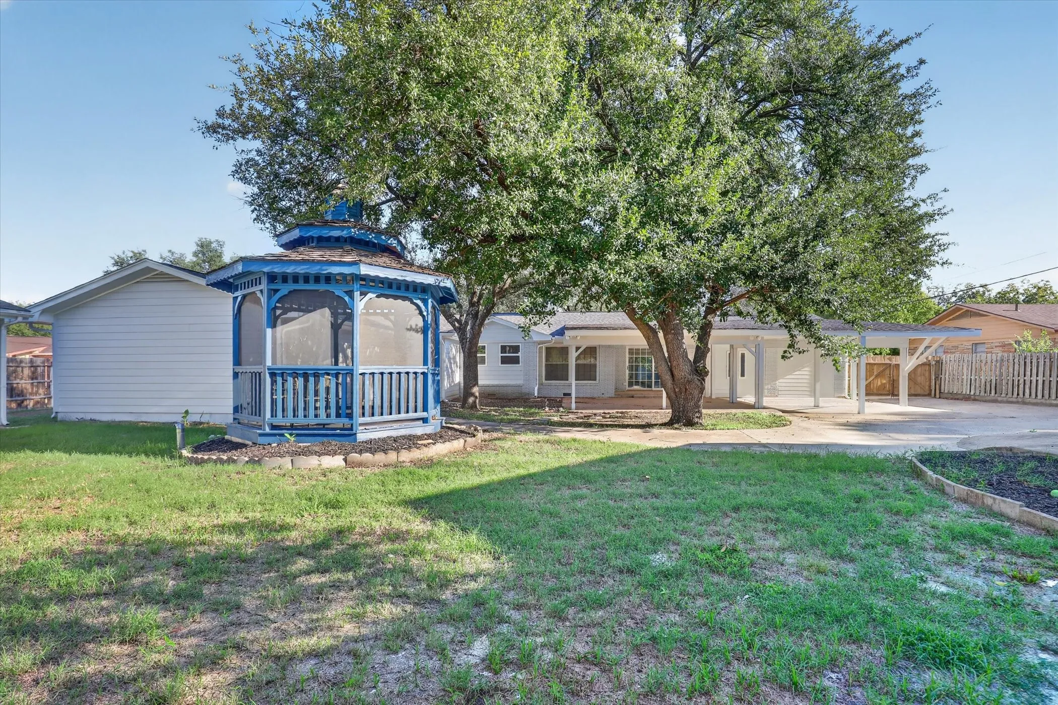 Expansive backyard featuring a blue gazebo, mature trees, and a covered carport