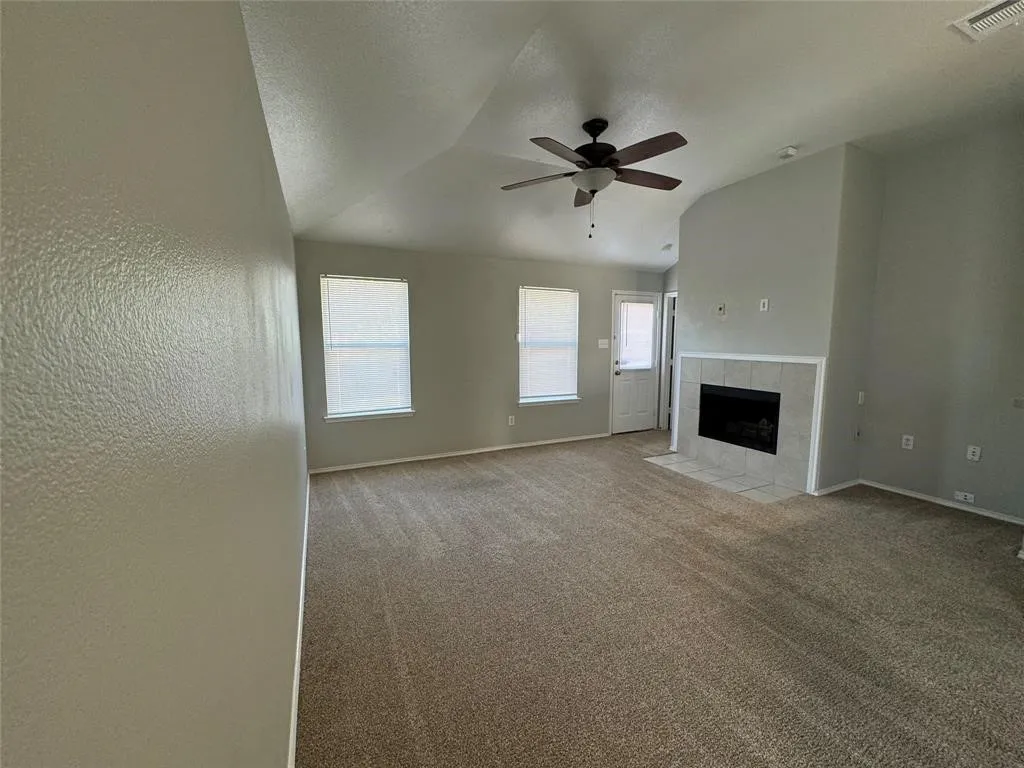 Vaulted ceiling with a ceiling fan, carpeted flooring, fireplace with a tiled surround, and two windows with blinds