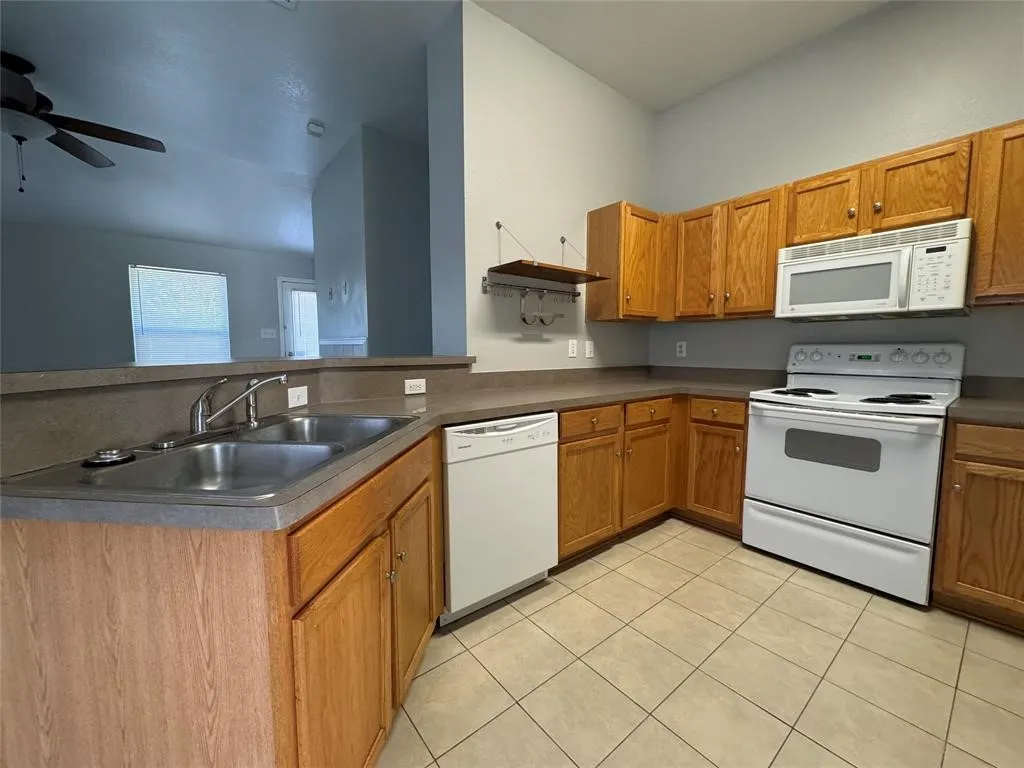Kitchen featuring wood cabinetry, solid surface countertops, a double basin stainless steel sink, tile flooring, and white appliances including a range, microwave, and dishwasher